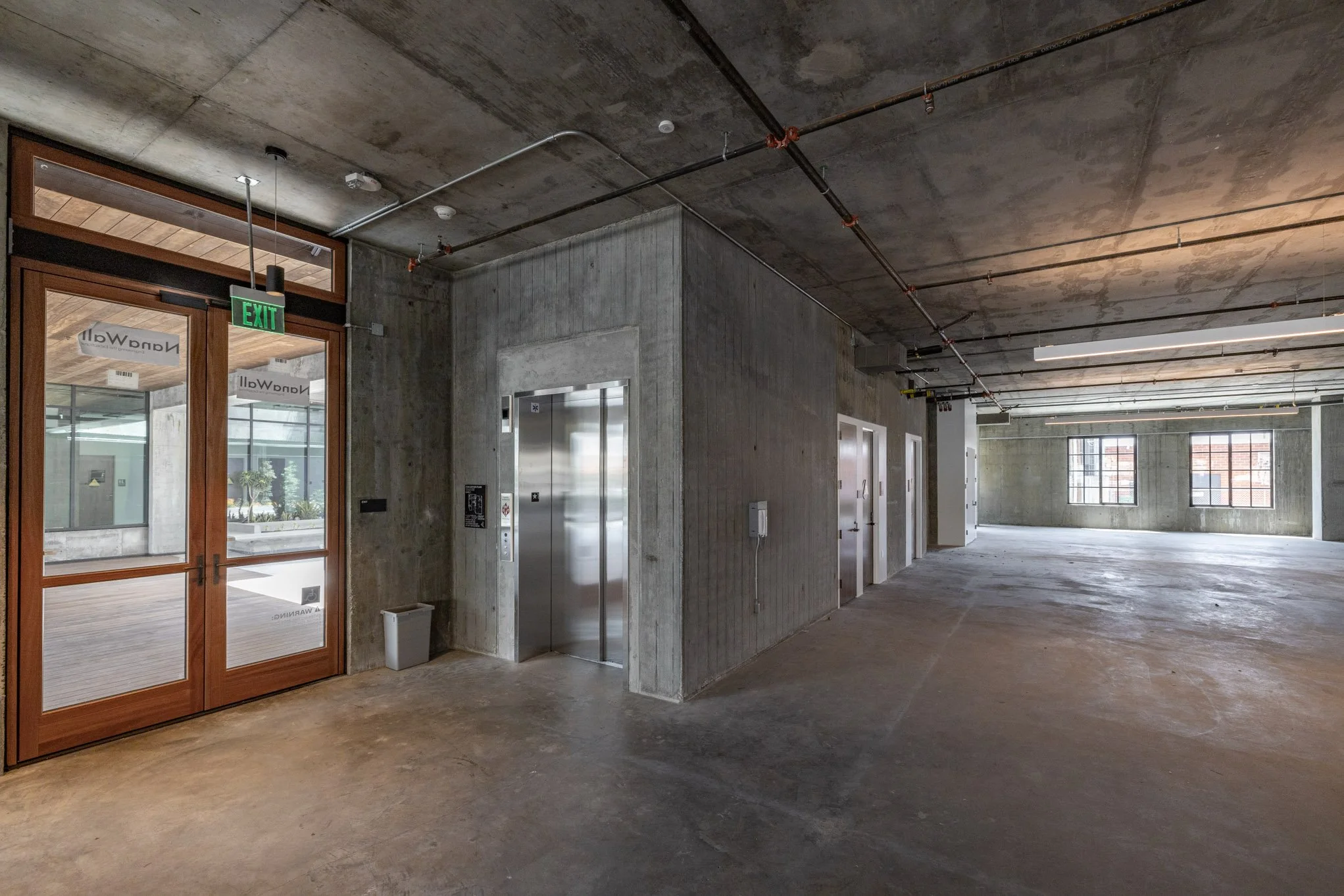 Empty industrial-style interior space with concrete walls and floor, multiple elevator doors, large windows, and exposed ceiling pipes.