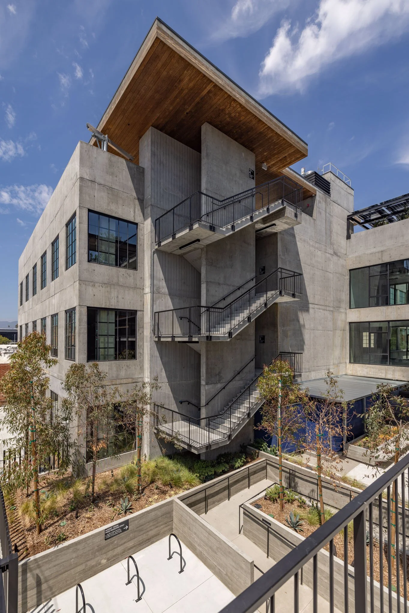 Modern concrete building with external staircases, large windows, and rooftop deck, surrounded by landscaped garden and young trees.