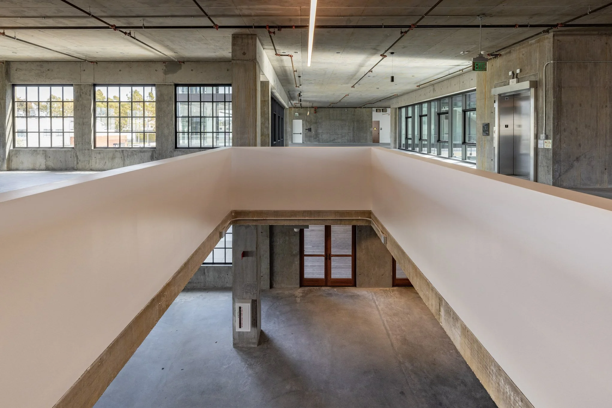 Empty modern interior with large windows, exposed concrete walls, an elevator, and an open staircase area in a building under construction or renovation.