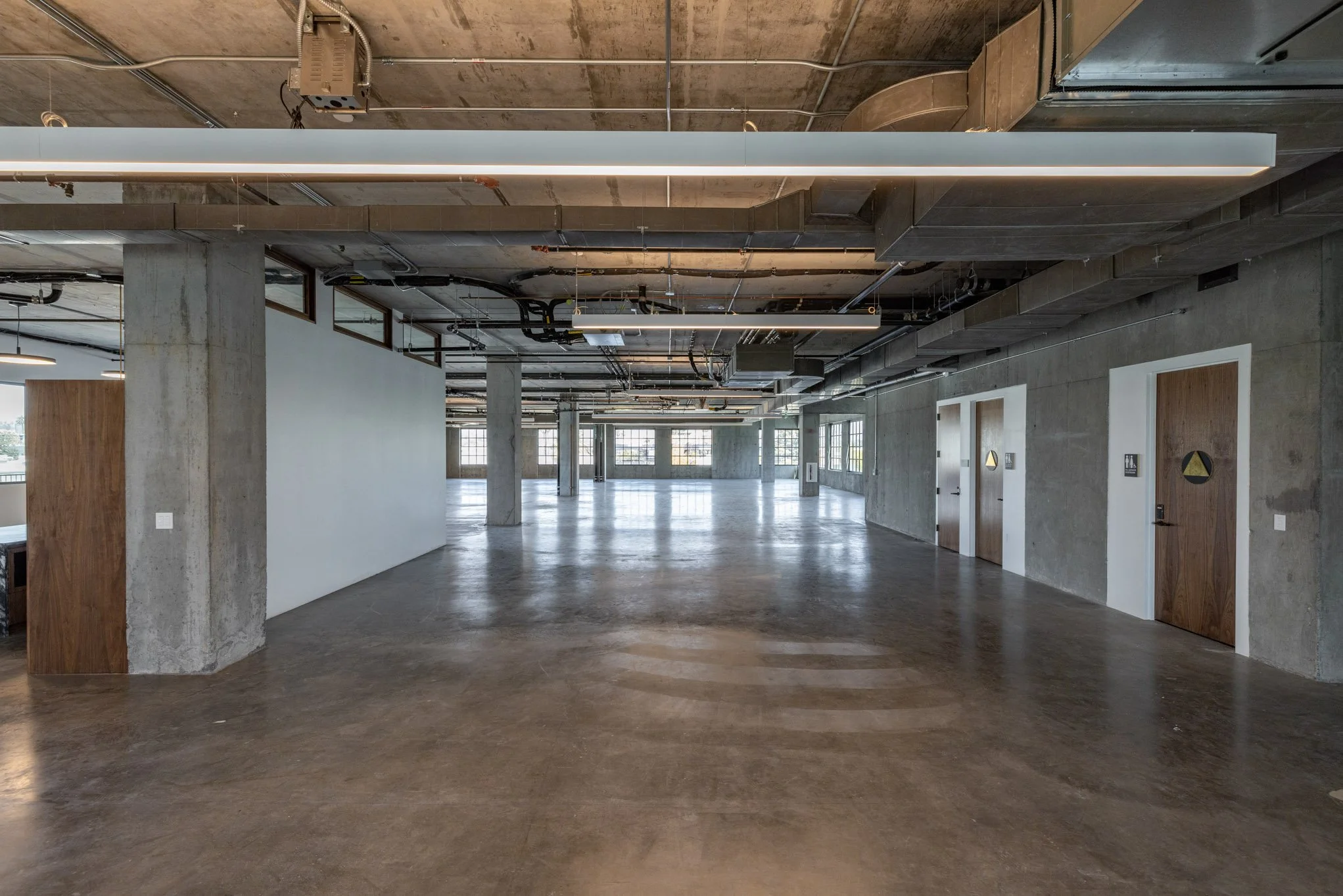 Empty industrial-style open space with concrete floors, exposed ceiling pipes, and multiple restroom doors along the right wall.