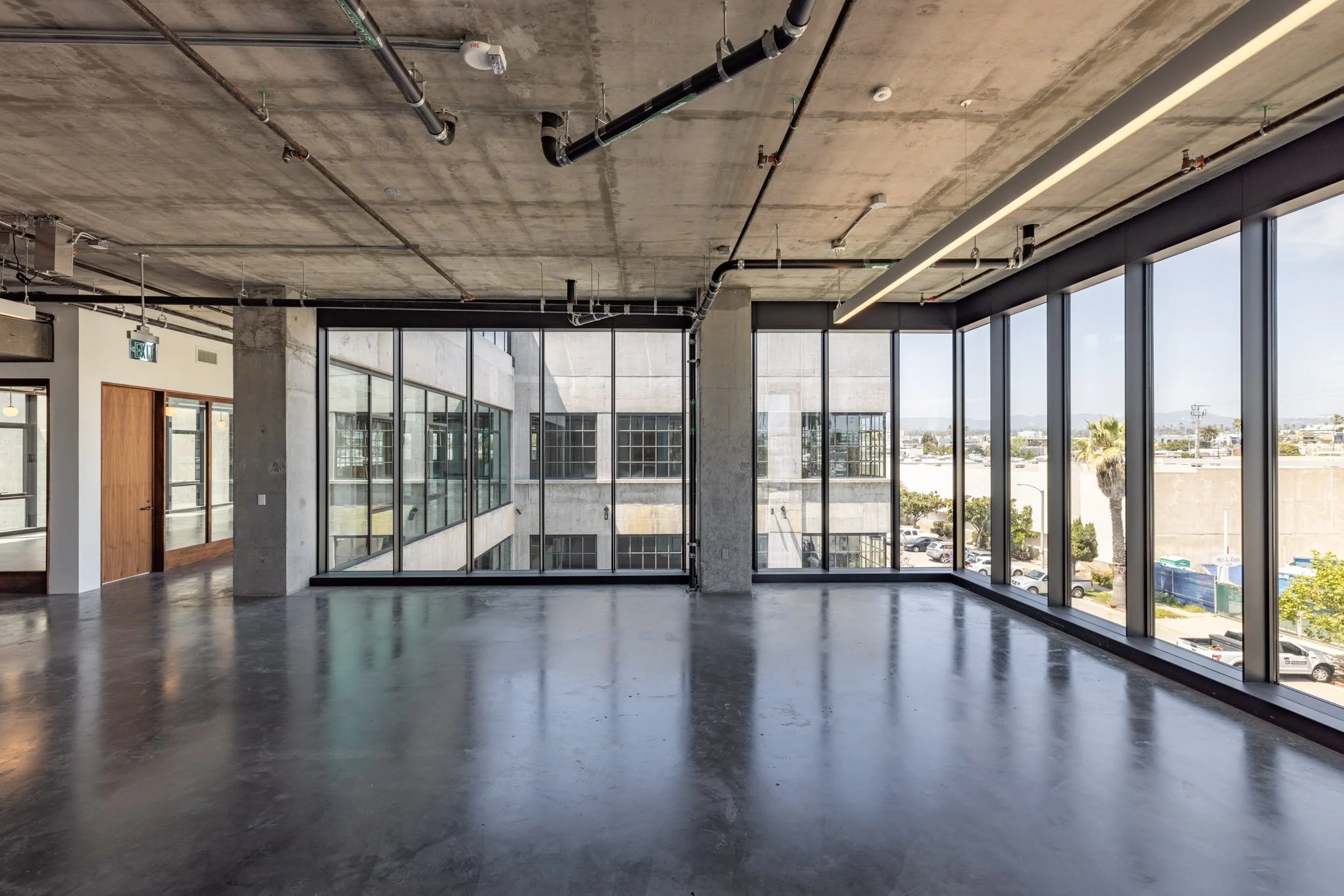 Empty modern office space with large floor-to-ceiling windows, polished concrete floors, exposed ceiling pipes, and a view of city buildings outside.