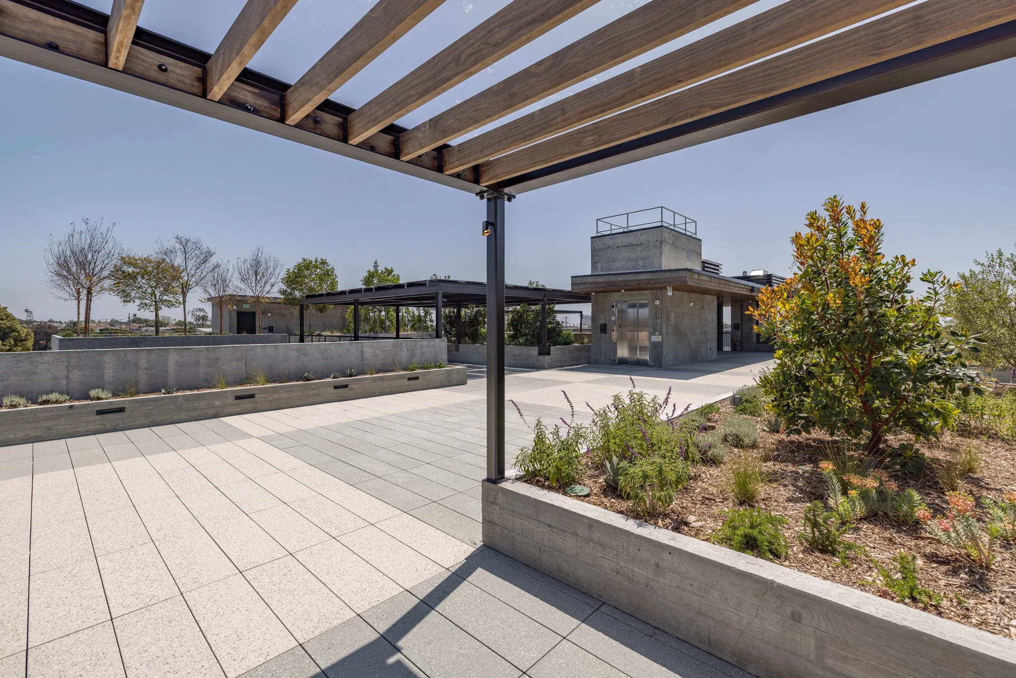 Modern outdoor terrace with tiled flooring, wooden pergola overhead, landscaped planter with greenery, and a concrete building in the background under clear blue skies.