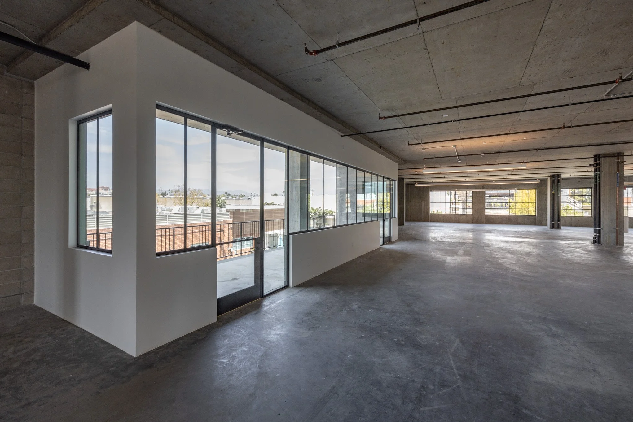 Empty office space with large windows, unfinished concrete floors and ceiling, and exposed piping.