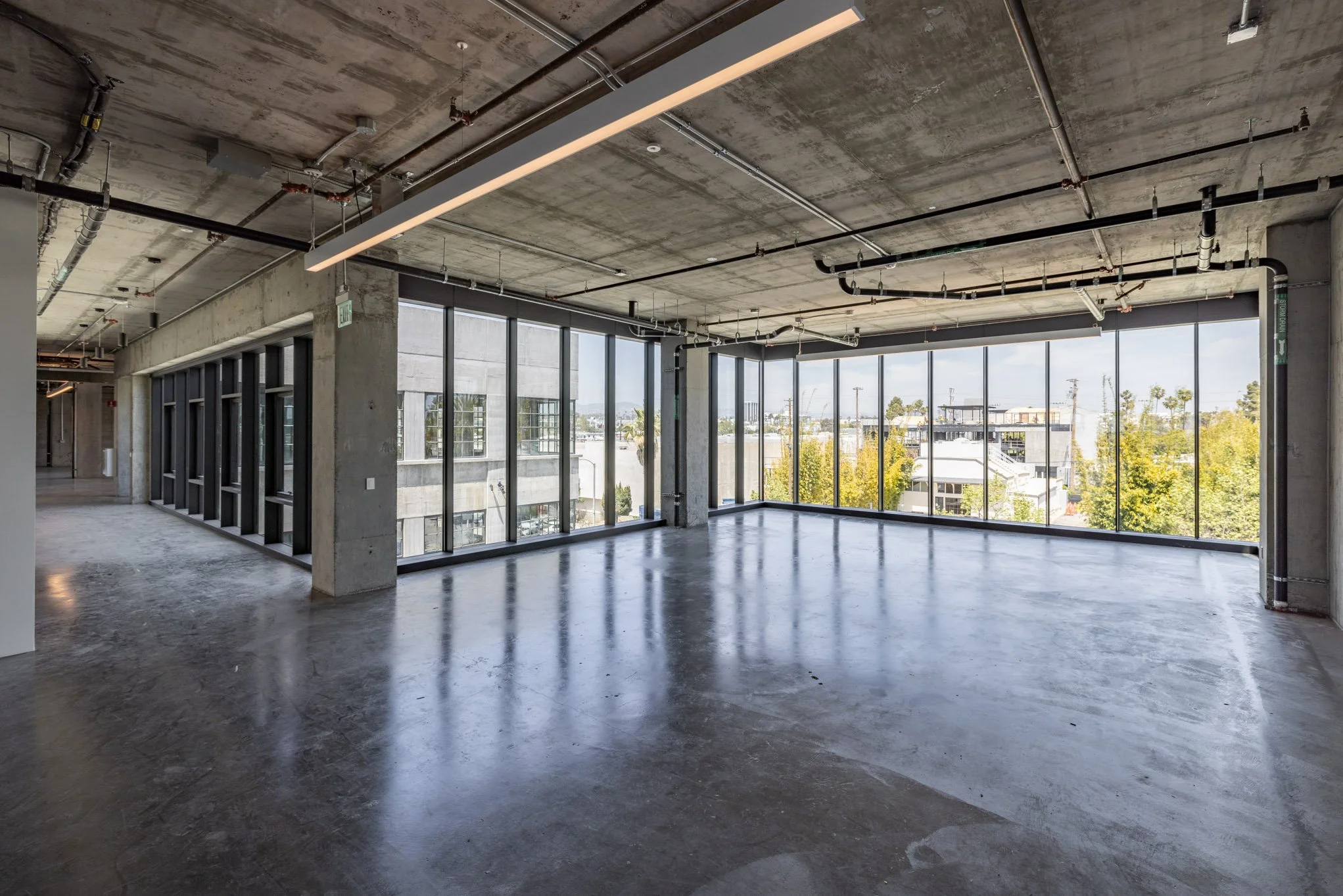 Empty modern office space with large floor-to-ceiling windows showing a city view, raw concrete floors and ceiling, visible ductwork, and a sleek linear ceiling light.