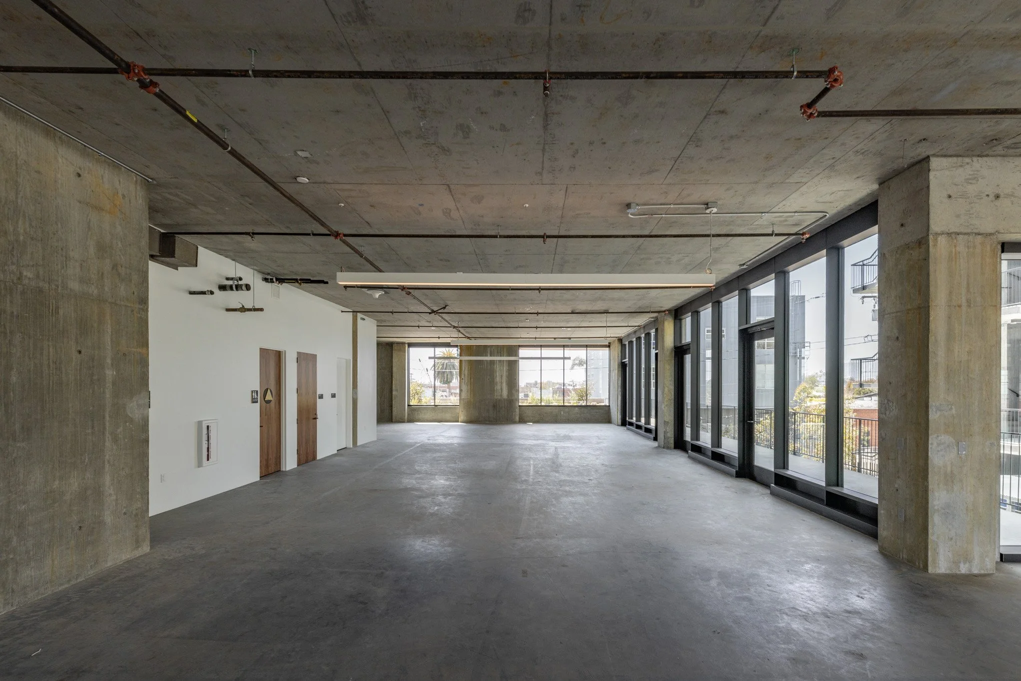 Empty modern commercial or office space with large glass windows, concrete walls and ceiling, and an unfinished floor.