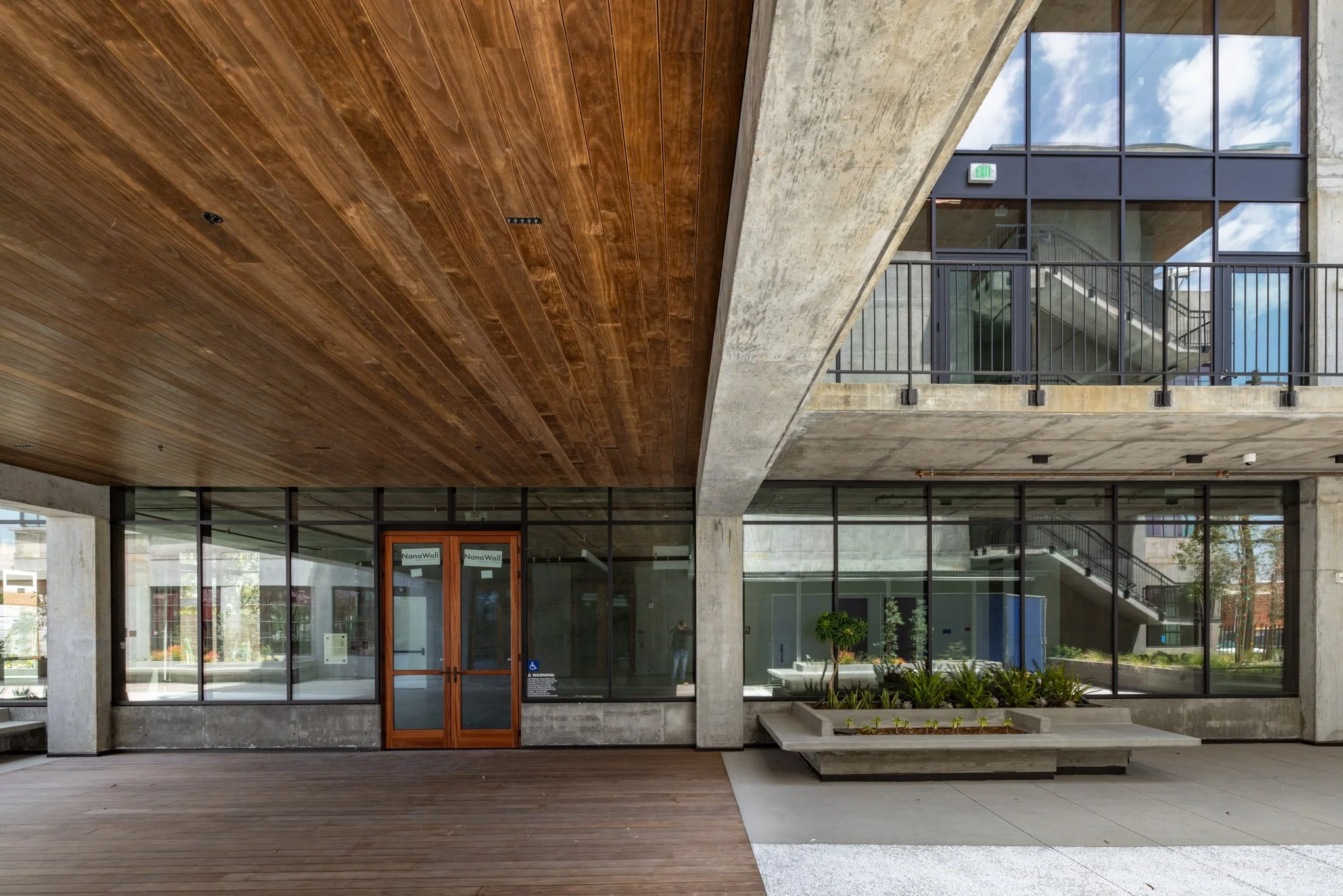 Modern building with glass walls, concrete columns, and wooden ceiling, featuring a small landscaped plant area and stairs visible through the windows.