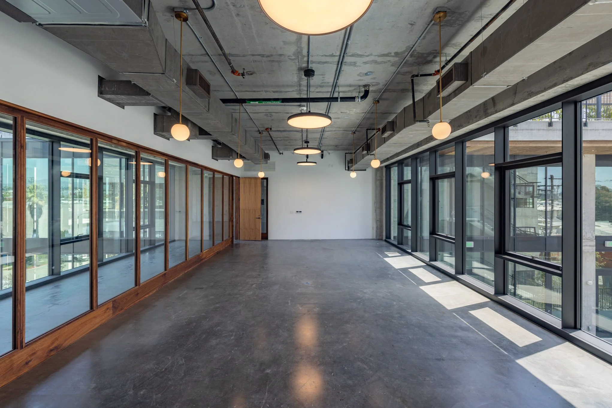 Empty modern office space with concrete ceiling, large floor-to-ceiling windows, and hanging round light fixtures.