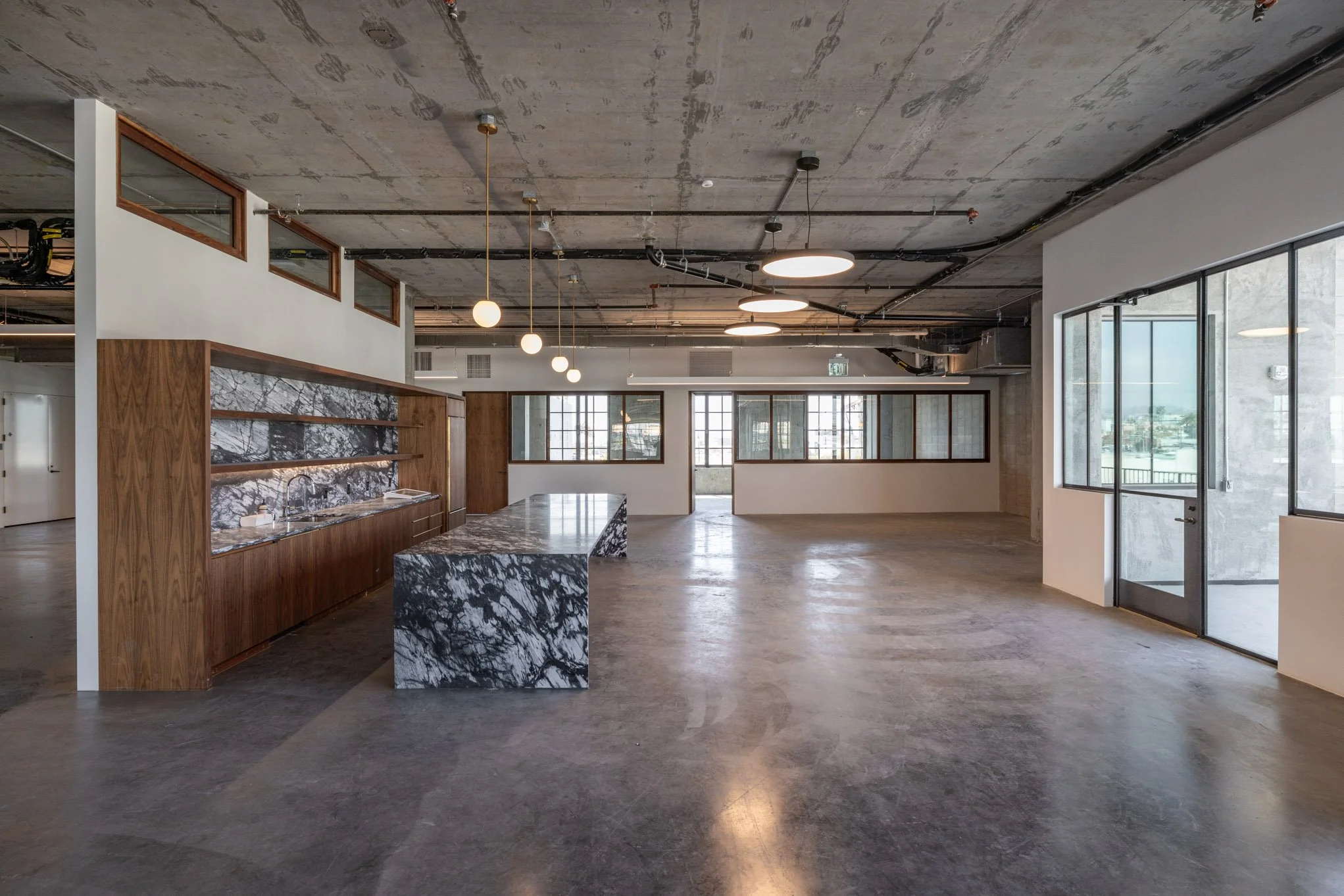 Empty industrial-style interior space with concrete floors, exposed ceiling with lighting fixtures, large windows, a marble kitchen island, and a built-in marble shelf with wooden accents.