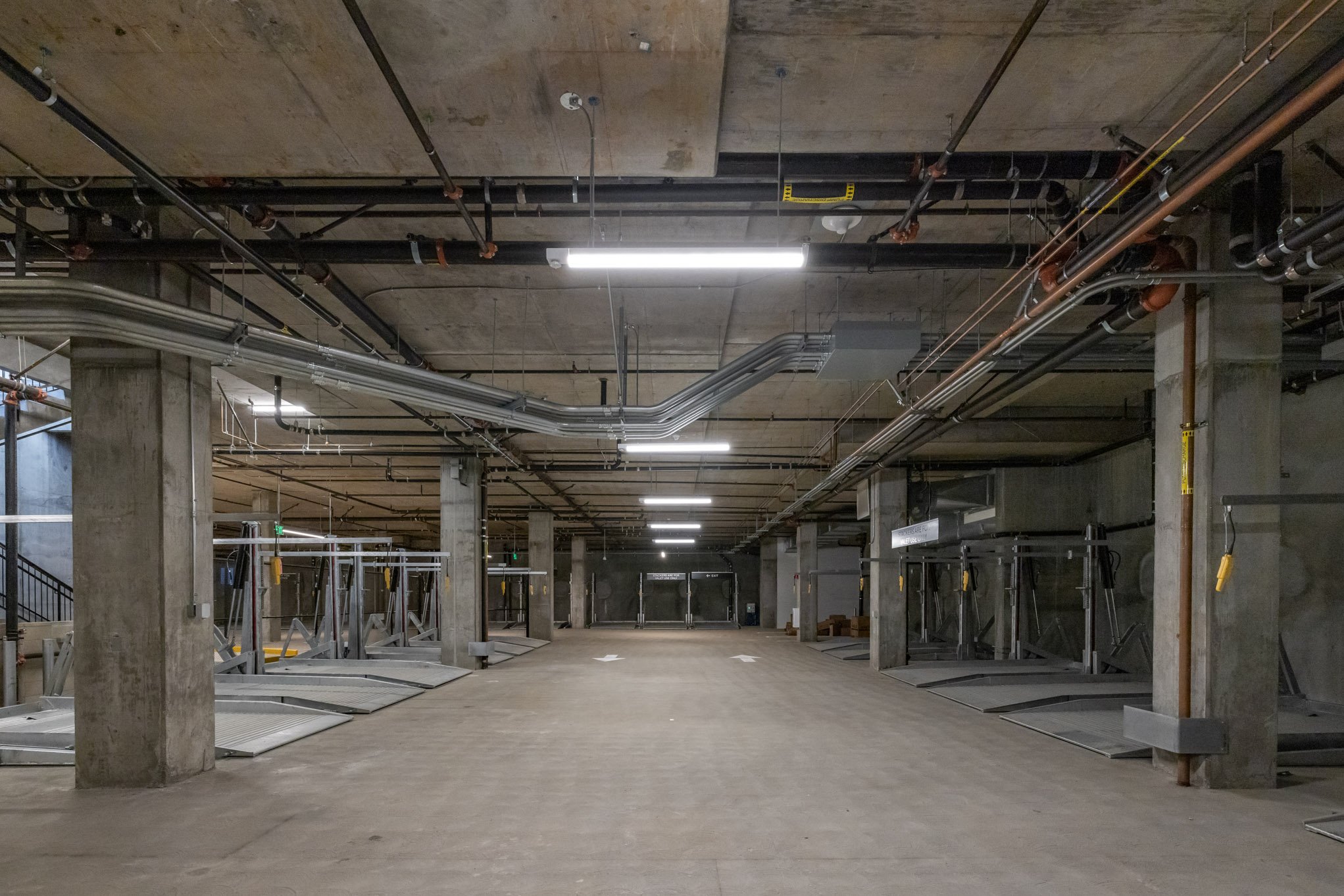 Empty indoor parking garage with concrete pillars, ceiling pipes, and bike racks, illuminated by overhead fluorescent lights.