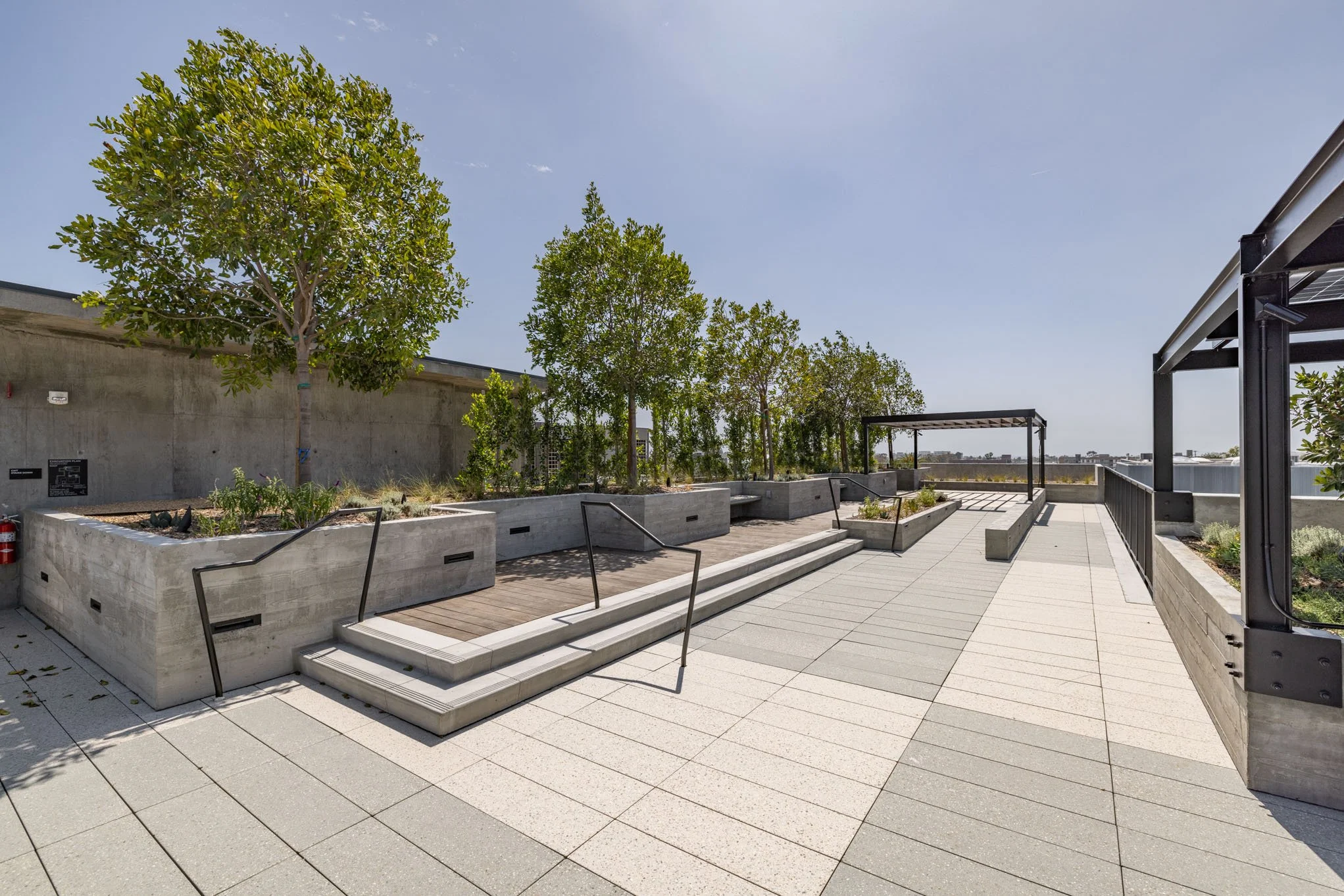 Rooftop terrace with trees, elevated garden beds, stairs, and seating areas under a clear blue sky.