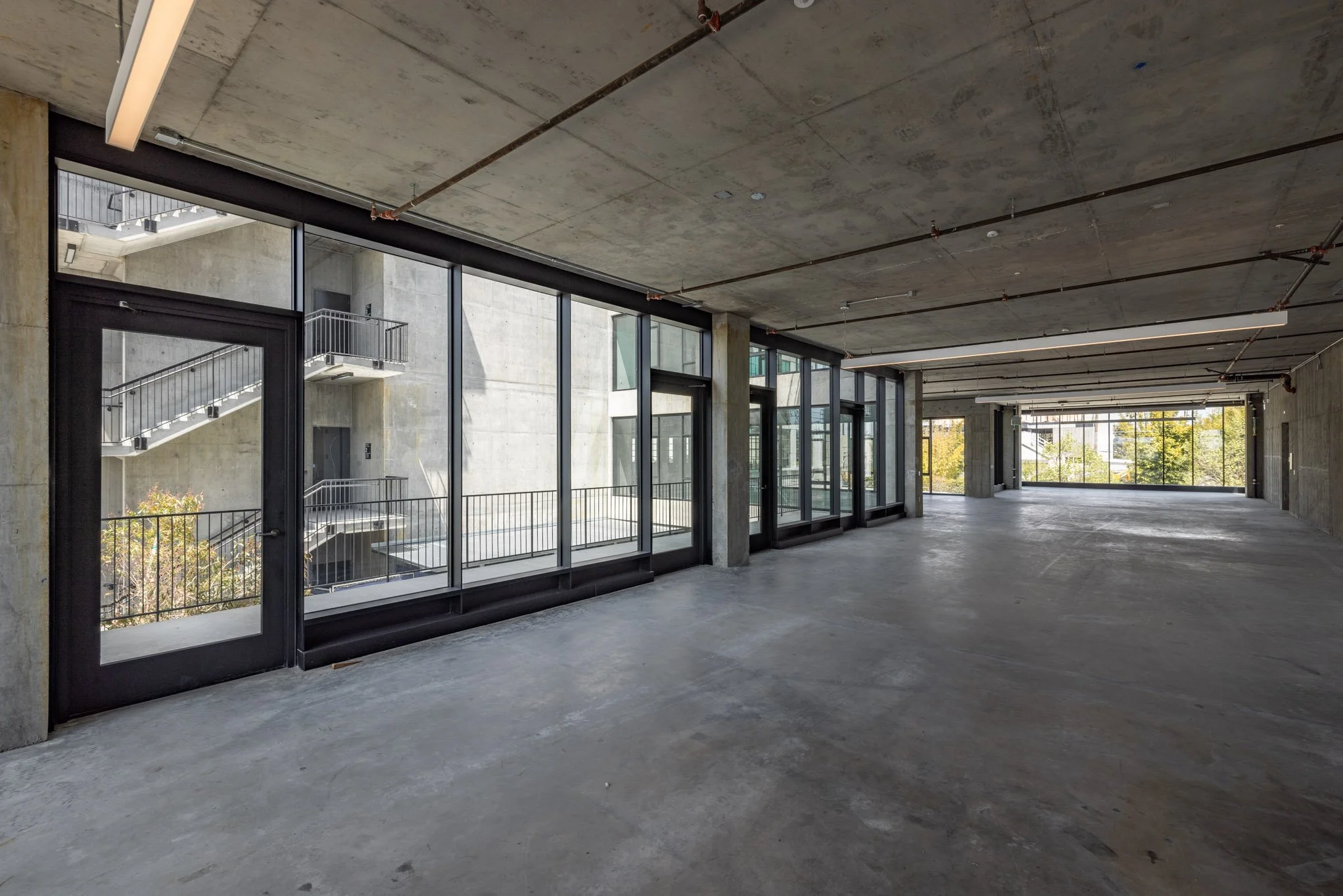 An empty, unfinished commercial interior space with concrete floors, exposed concrete ceiling with visible pipes, large floor-to-ceiling windows along one wall, and a door leading to an outdoor fire escape staircase.