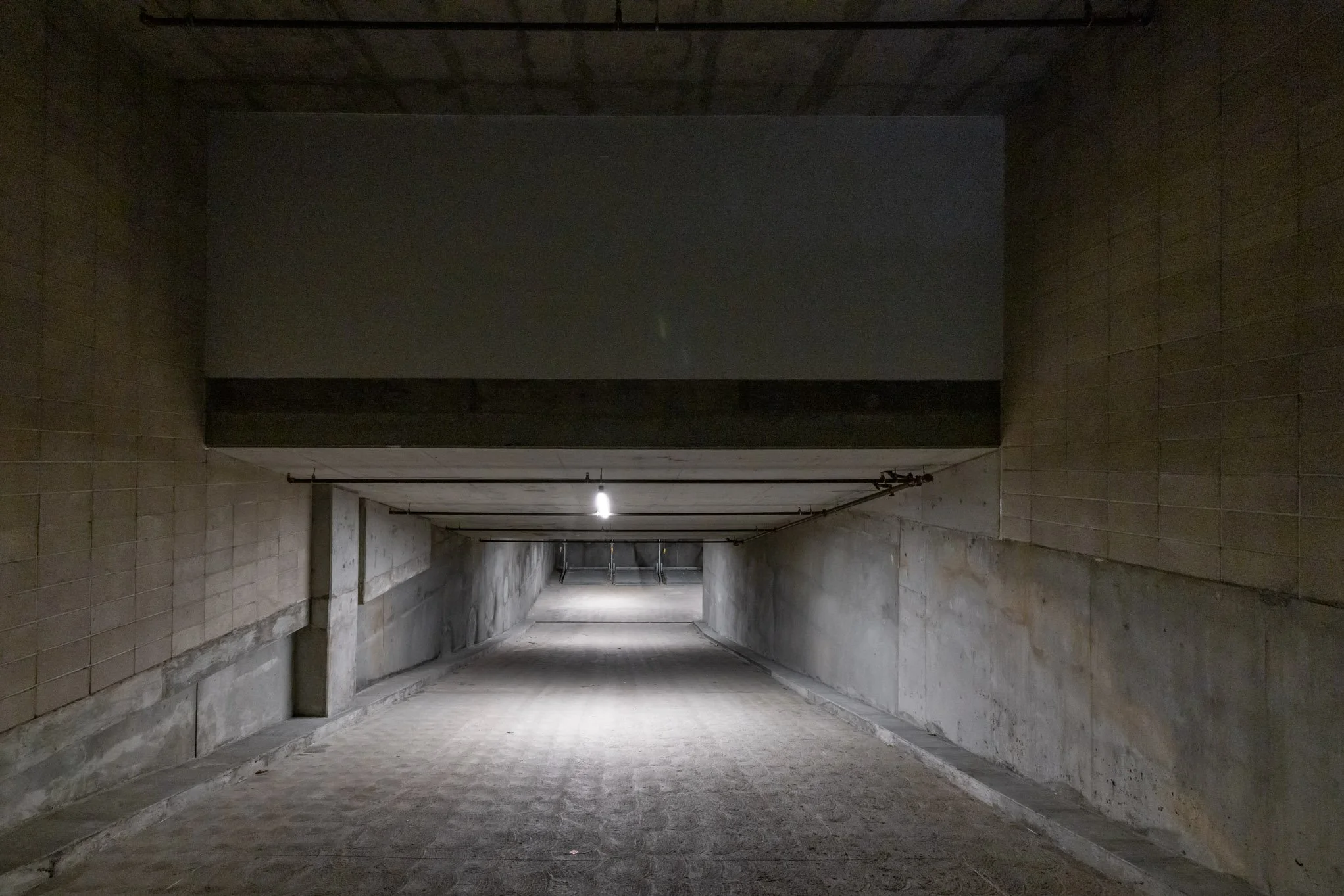 Underground parking garage with concrete walls and ceiling, dim lighting, and a short tunnel leading to an exit or open area.