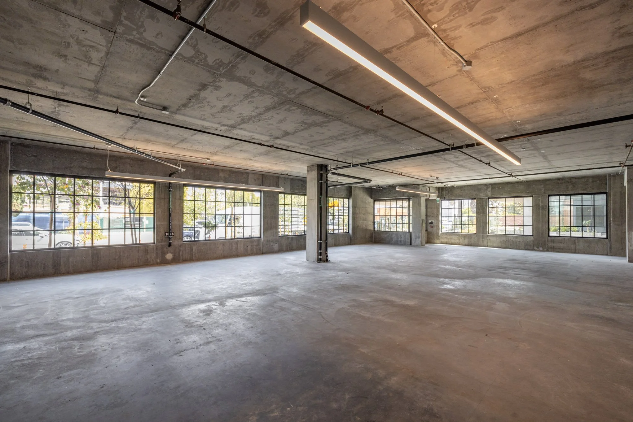 Empty commercial space with large windows, unfinished concrete floors, exposed ceiling with hanging lights, and visible electrical conduits.