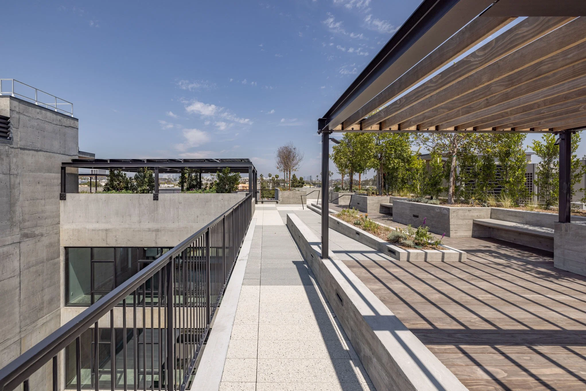 Modern rooftop terrace with concrete planters, wooden benches, trees, and a metal pergola under a partly cloudy sky.