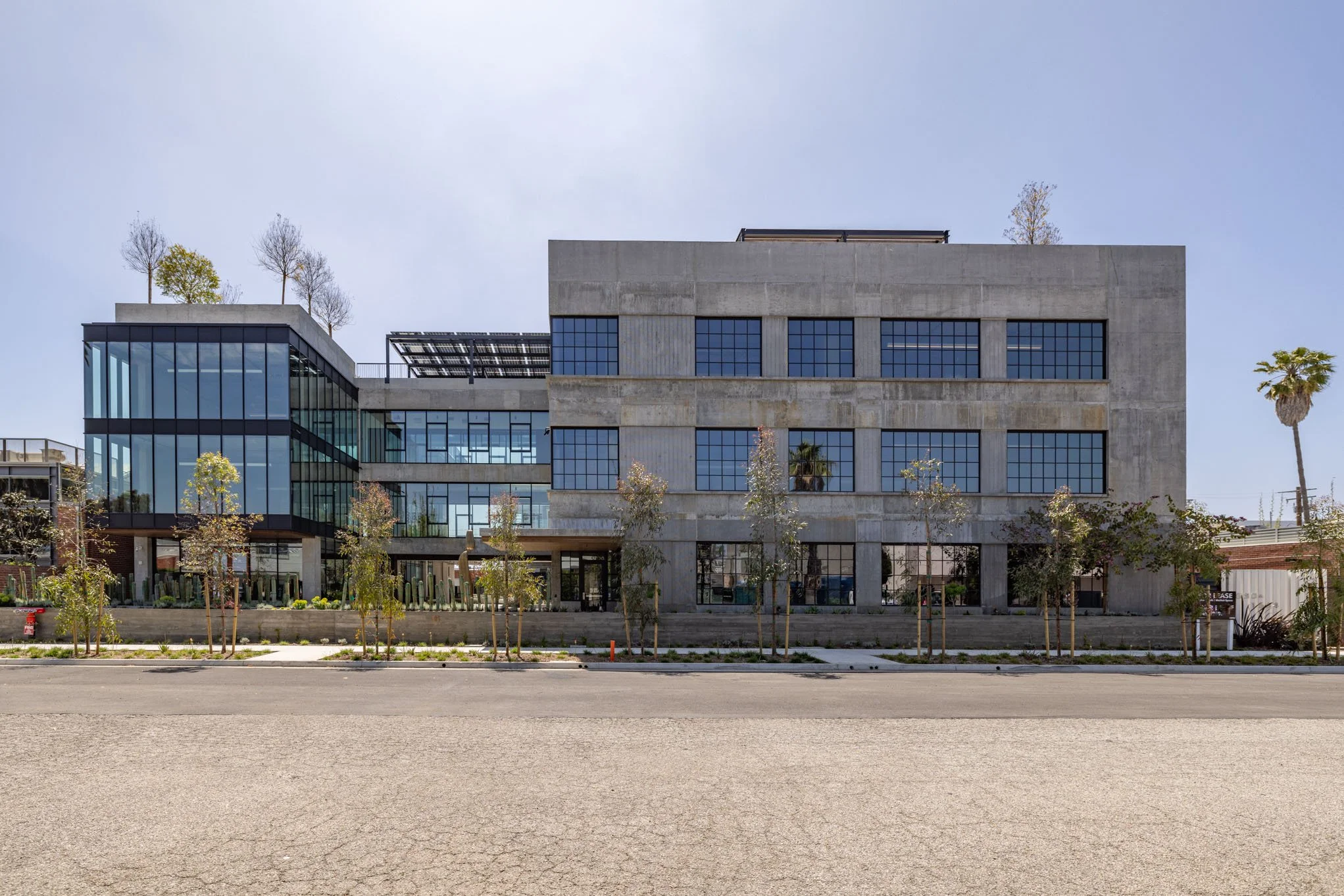 Modern multi-story building with large glass windows and concrete exterior, surrounded by small trees and a clear blue sky.