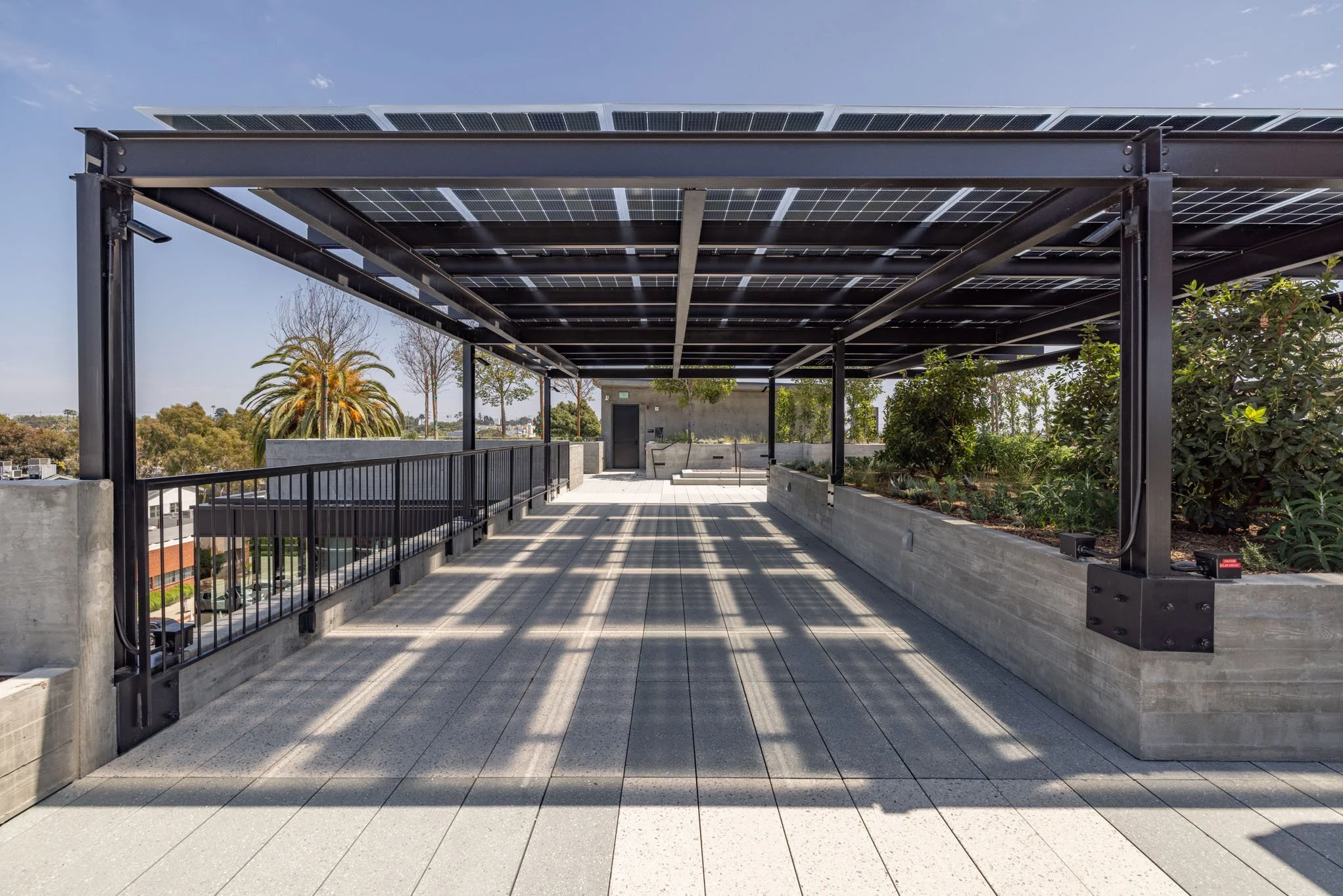 Rooftop terrace with solar-paneled pergola, concrete planters with greenery, and a urban view in the background.