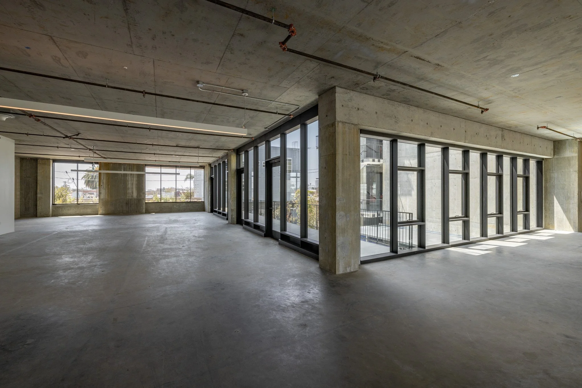 Empty unfinished interior space with concrete floors, walls, and ceiling, large windows with black frames, and exposed piping on the ceiling.