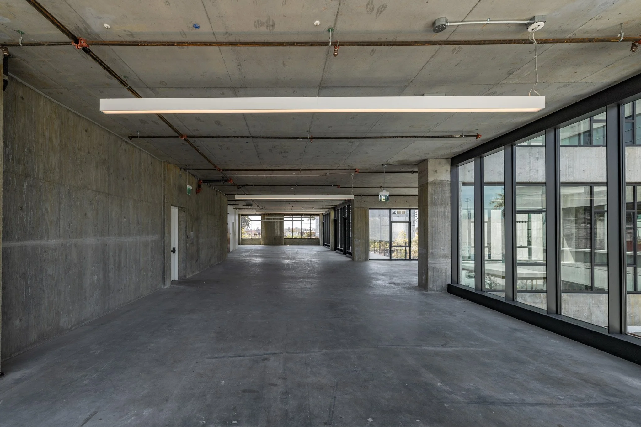 Empty modern commercial space with concrete walls, large floor-to-ceiling windows, and concrete ceiling with hanging lights.