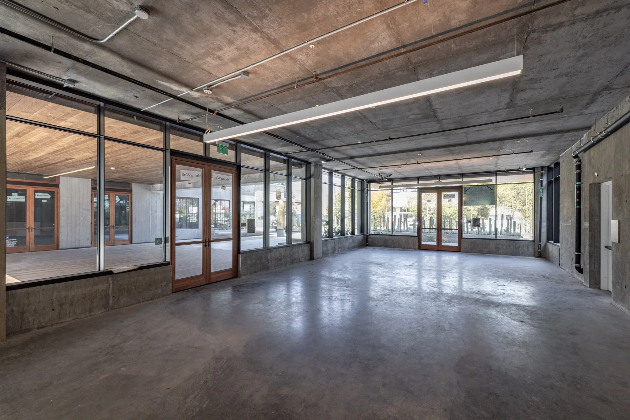 Empty commercial space with concrete floors, large front windows, and exposed ceiling pipes and lighting.