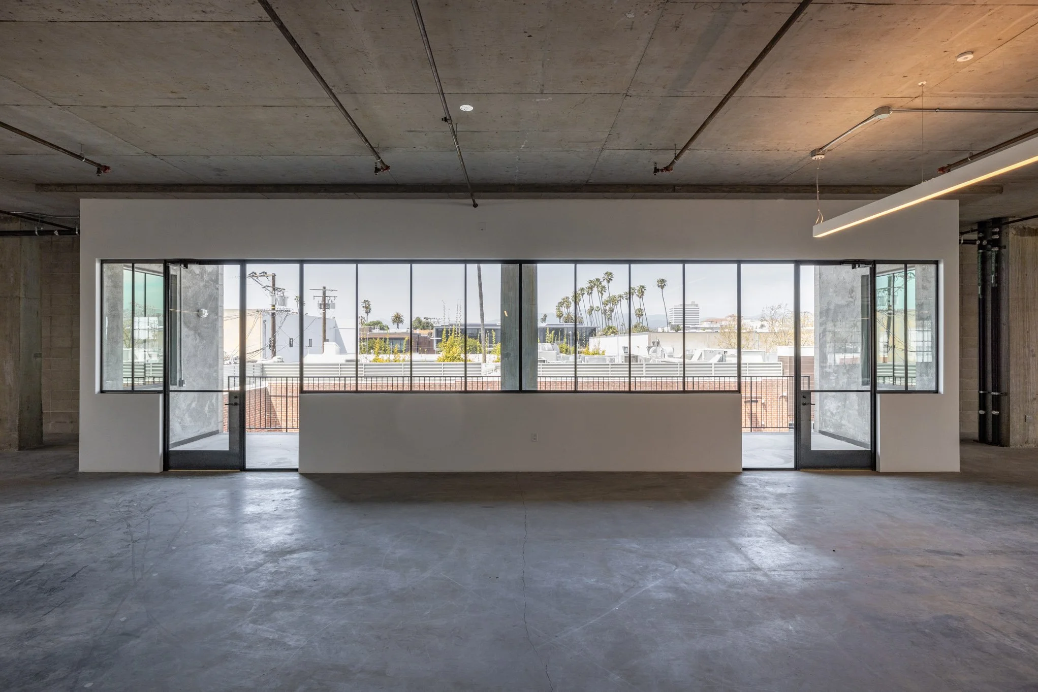 Empty industrial-style space with large windows and sliding glass doors leading to a balcony, overlooking palm trees and city buildings in the distance.