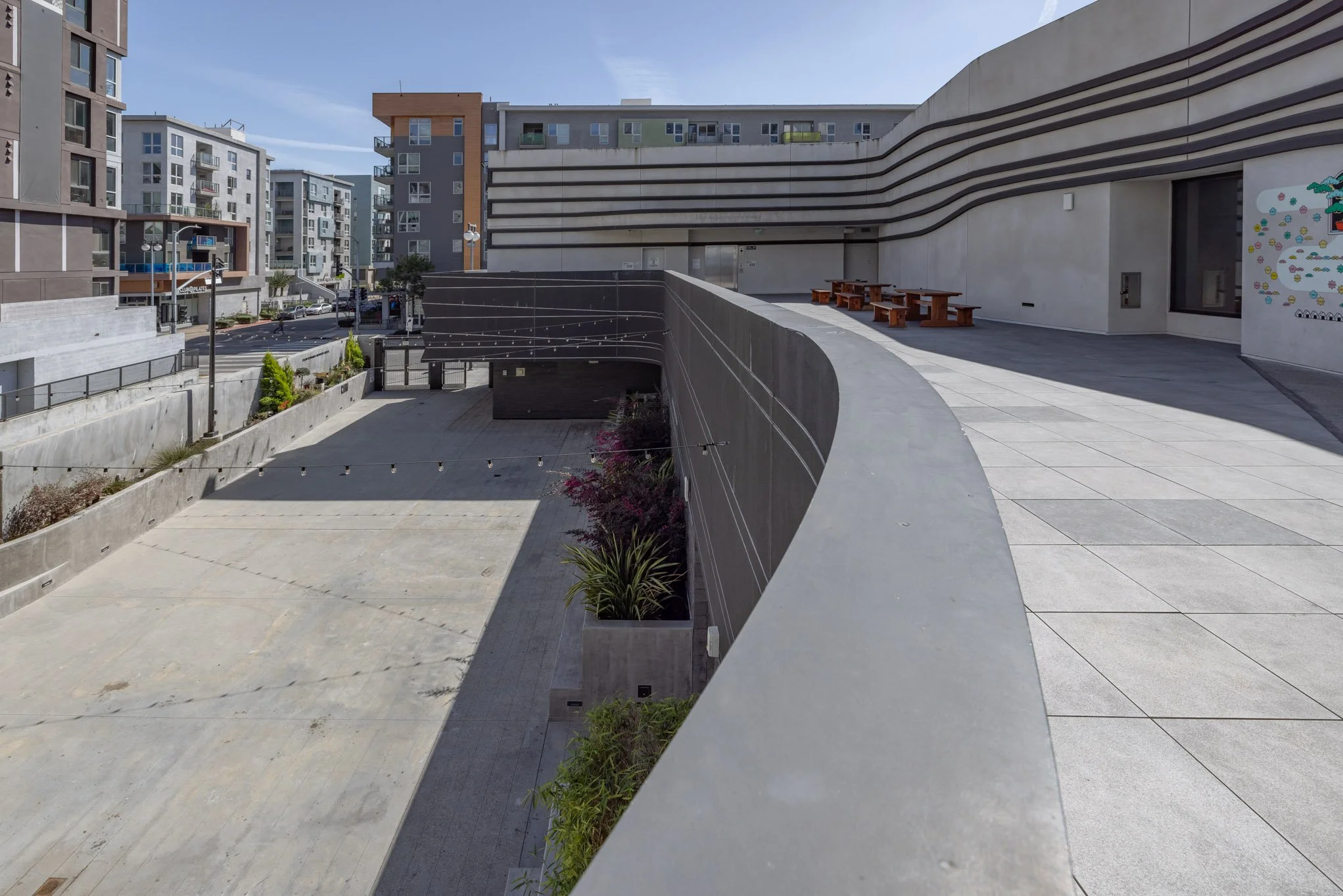 An outdoor urban space with modern apartment buildings, a concrete courtyard, and a rooftop seating area with picnic tables. There are string lights hanging and some plants along the edge.