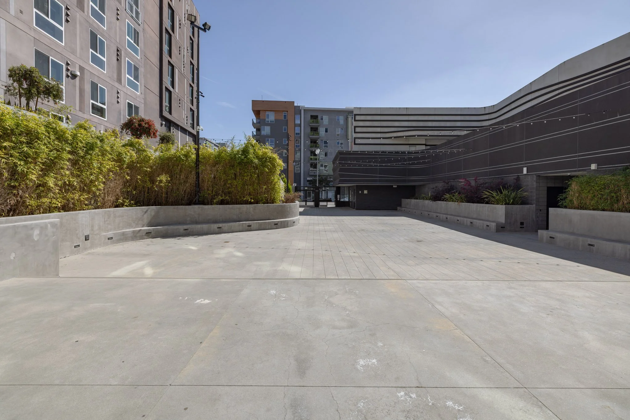 Empty concrete courtyard surrounded by modern apartment buildings with potted plants and string lights under a clear blue sky.