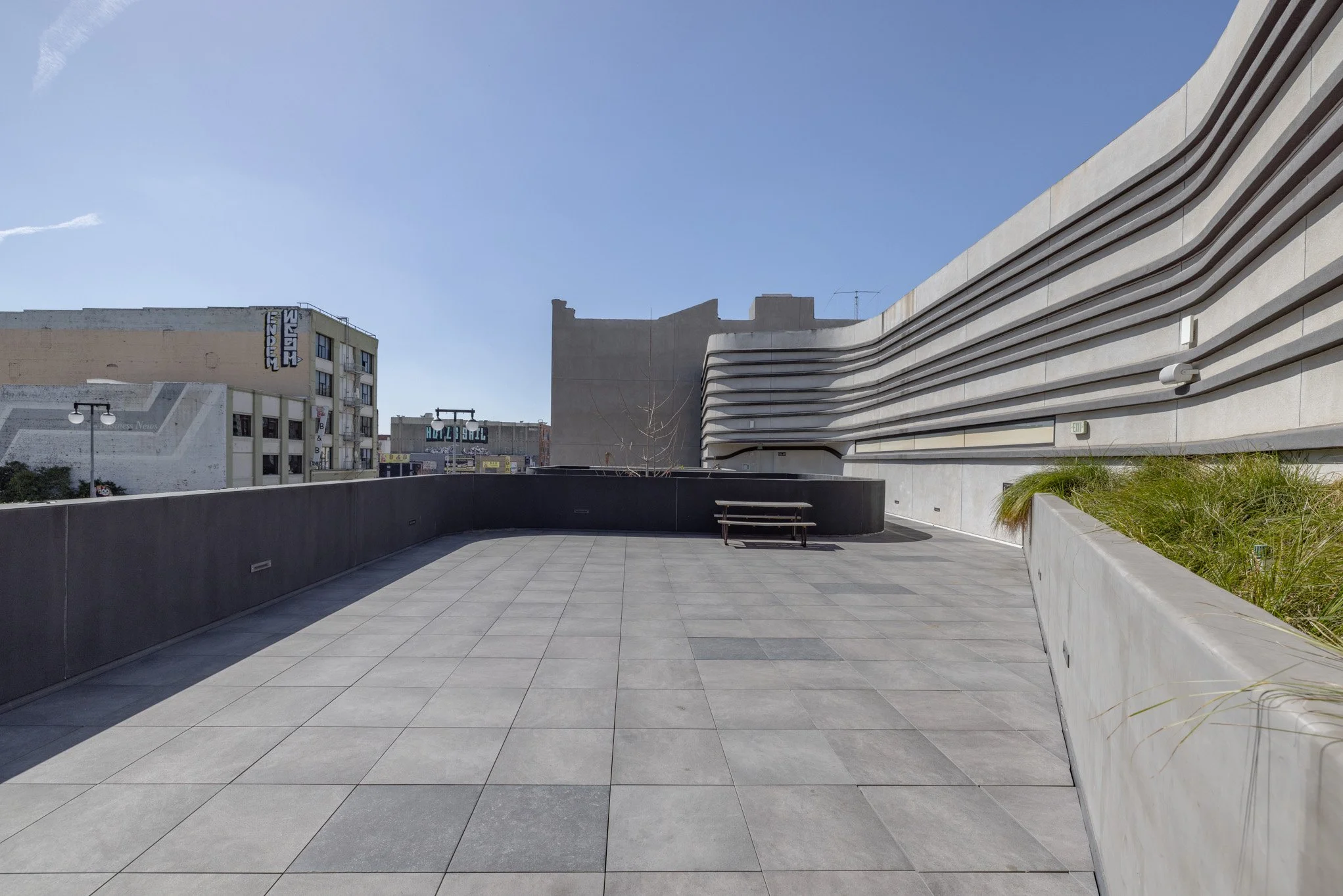 A spacious, modern outdoor terrace with tiled floor, a black concrete wall, and a bench, adjacent to a contemporary building with horizontal siding, in an urban area during daytime.