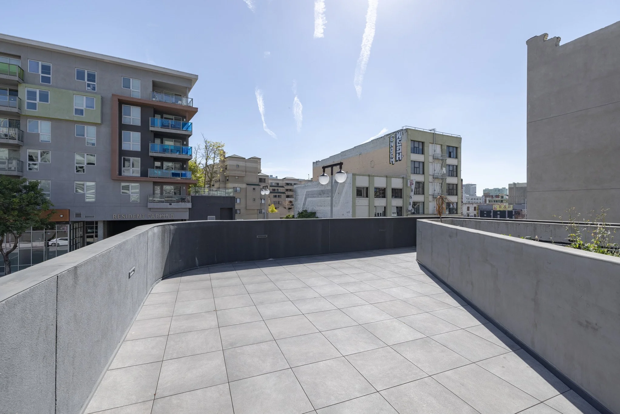 View of a rooftop terrace with light gray tiles, concrete railings, and city buildings in the background under a blue sky with wispy clouds.