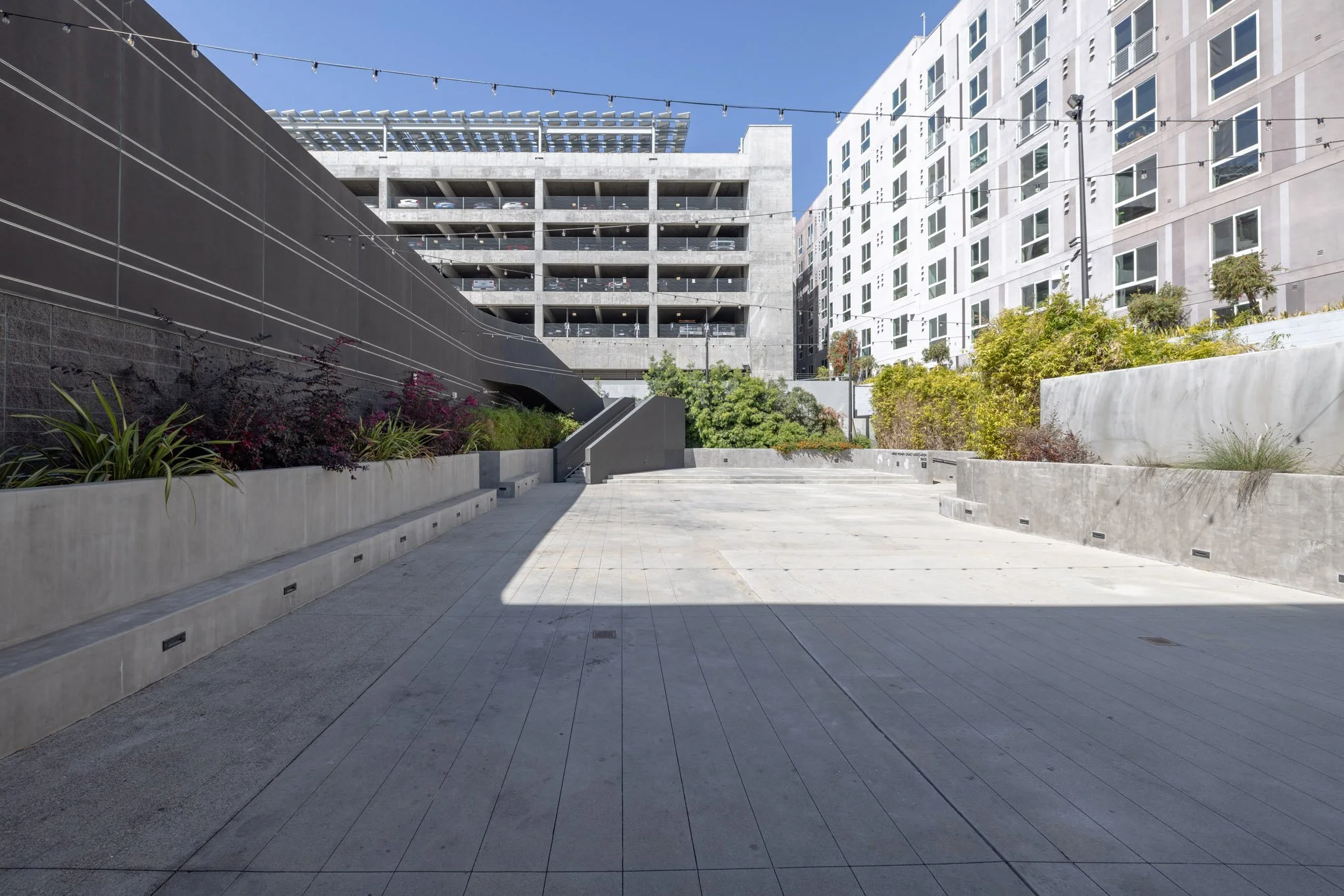 Empty urban outdoor space with concrete paving, planters with greenery, a staircase, and modern buildings in the background.