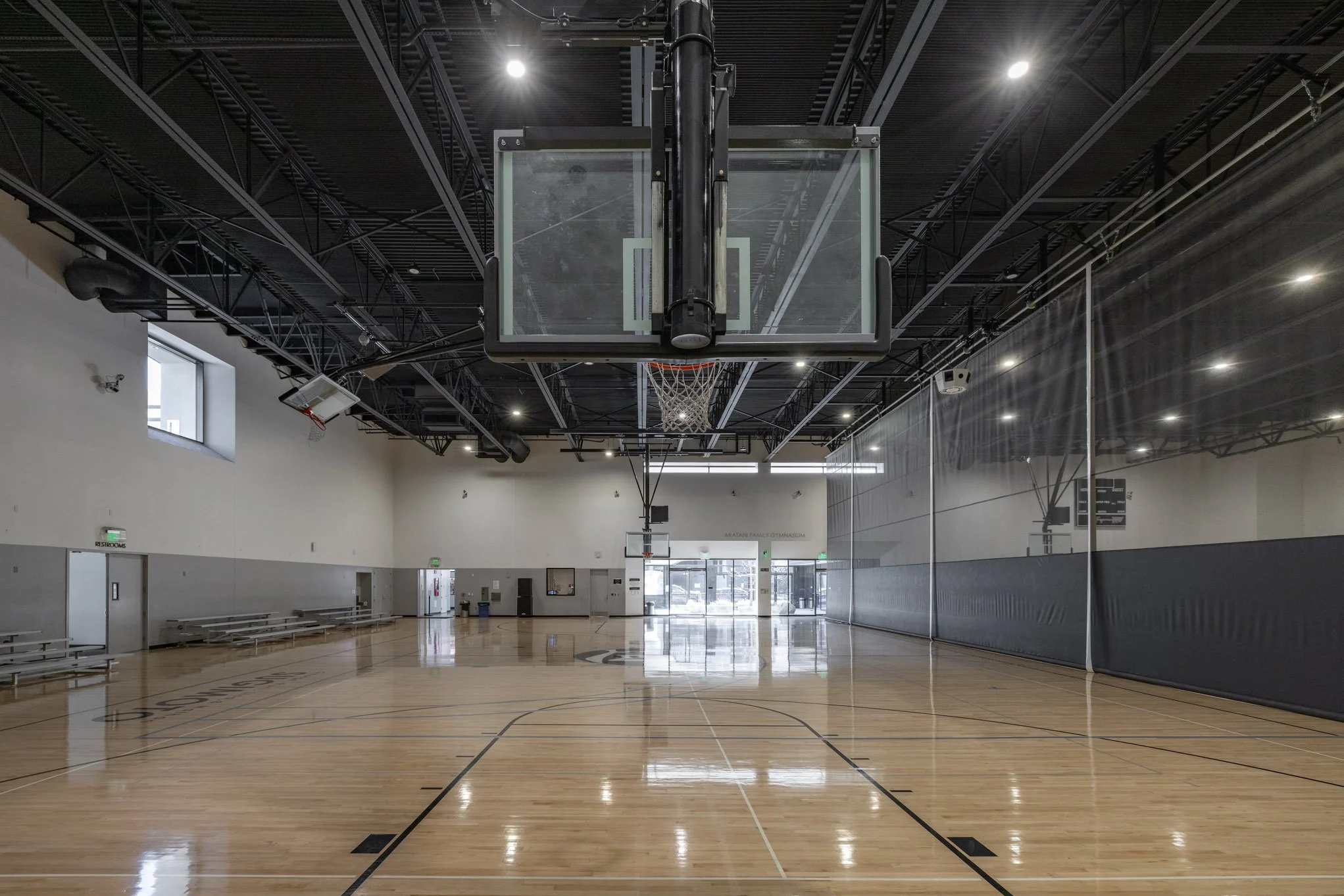 Empty indoor basketball court with hardwood flooring, basketball hoop, and glass backboard, surrounded by high ceiling and walls with windows and mirrors.