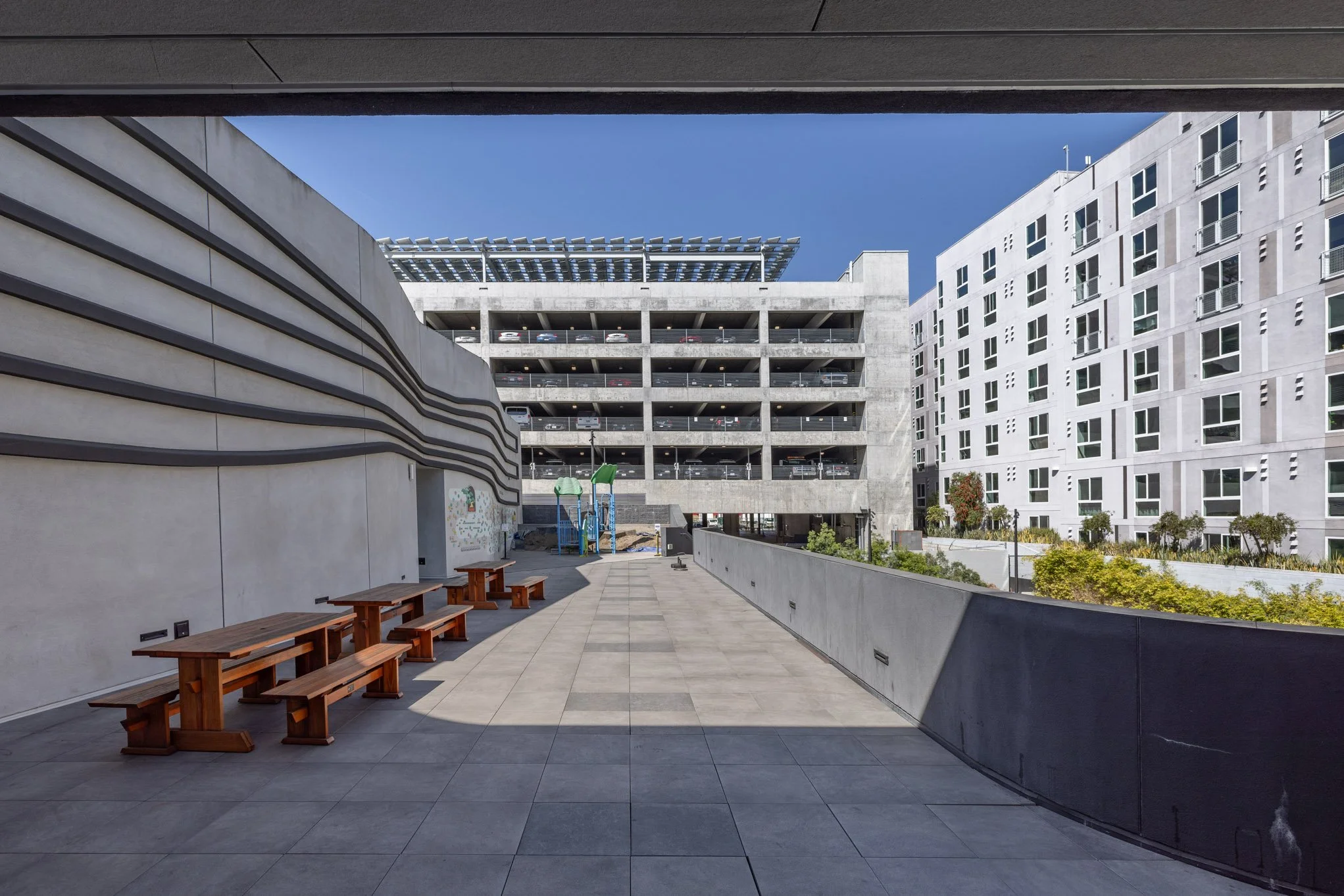 A modern urban courtyard with wooden benches, a multilevel parking garage, and a white residential building under a clear blue sky.