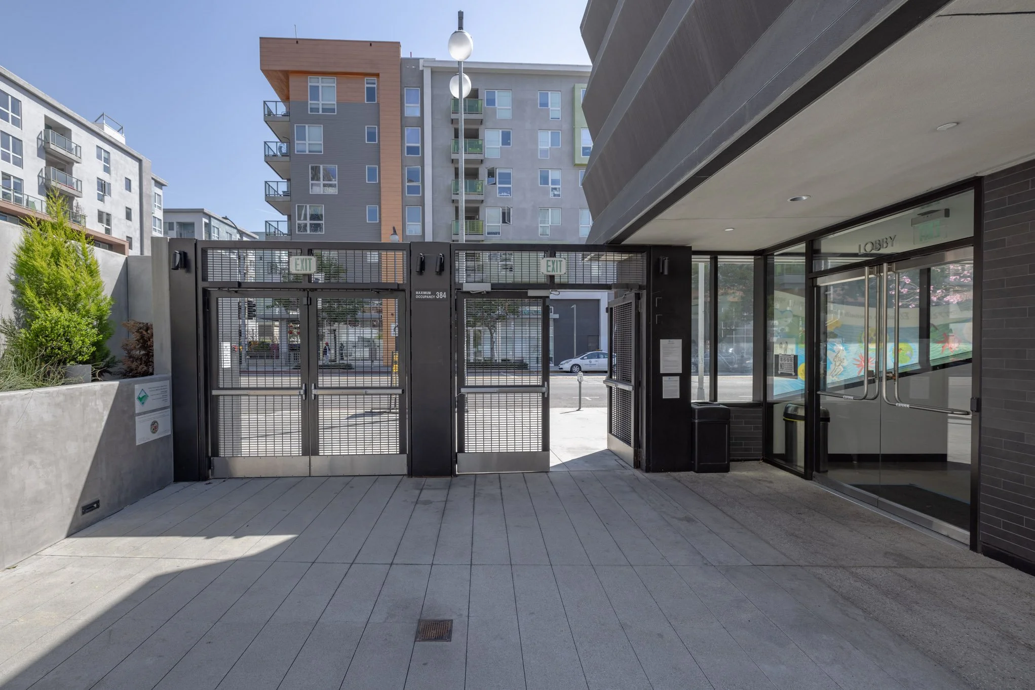 Entrance gate and lobby area of a modern apartment complex with tall residential buildings in the background, glass doors, and a sidewalk.