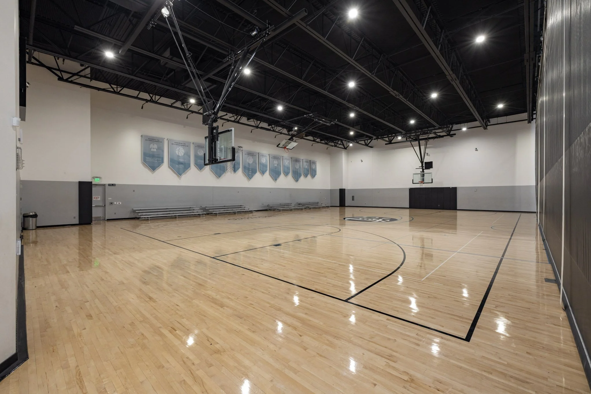 Empty indoor basketball court with wooden floor, basketball hoops, and bleachers, illuminated by ceiling lights.