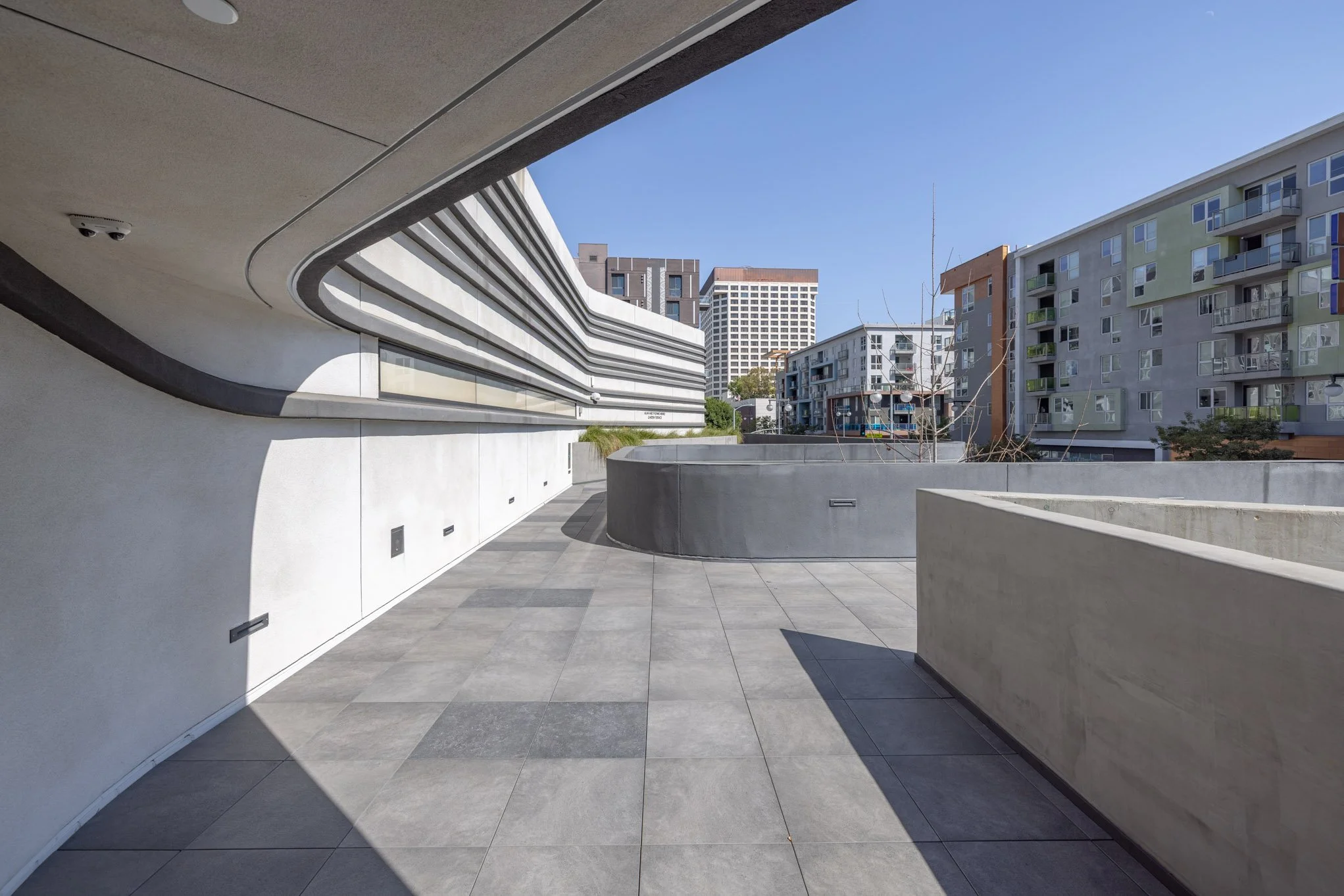 Empty modern urban balcony with curved concrete walls and gray tiled floor, overlooking surrounding buildings with balconies under clear blue sky.