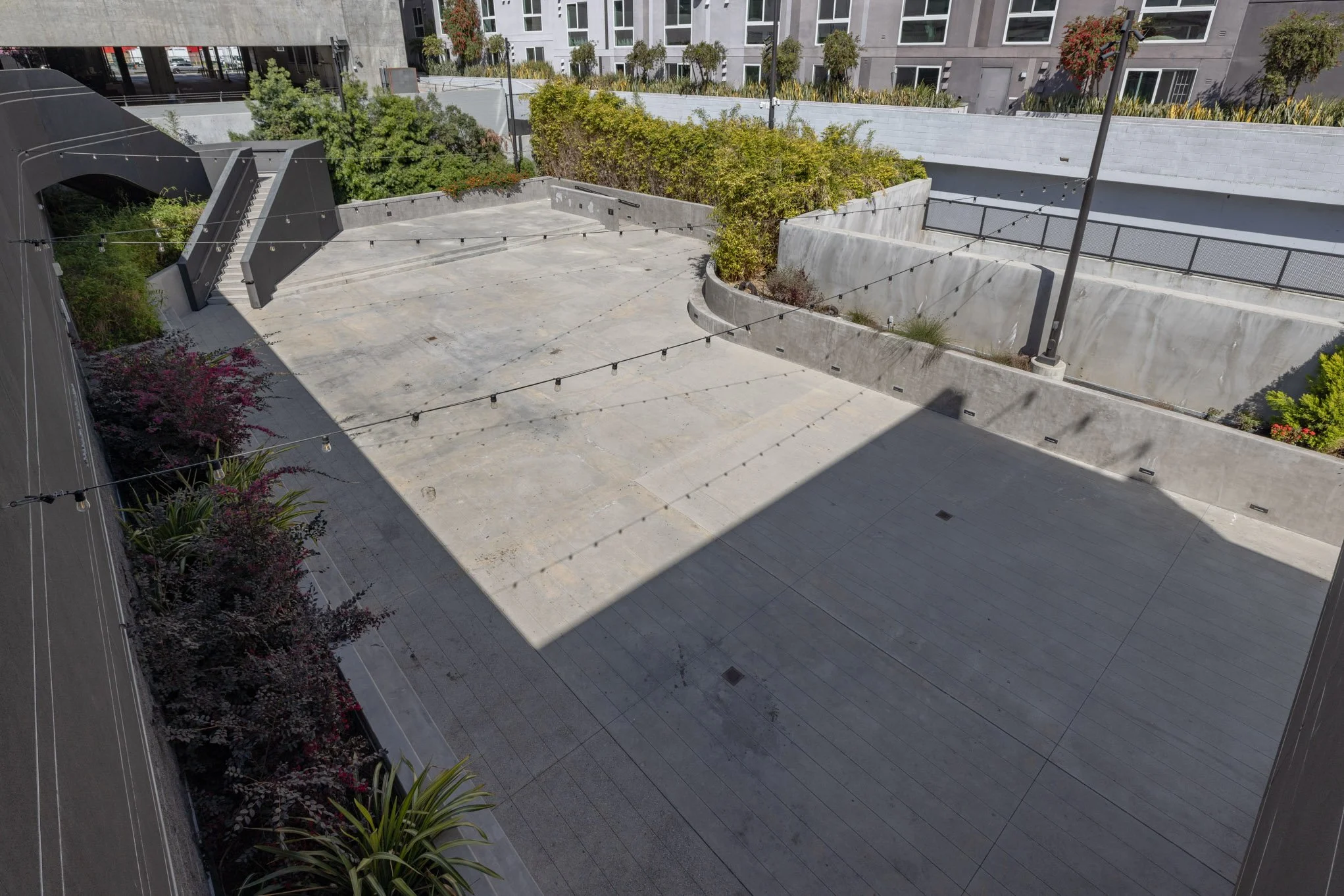 Empty outdoor urban courtyard with concrete surface, string lights, stairs, and surrounding landscaping, with apartment buildings in the background.