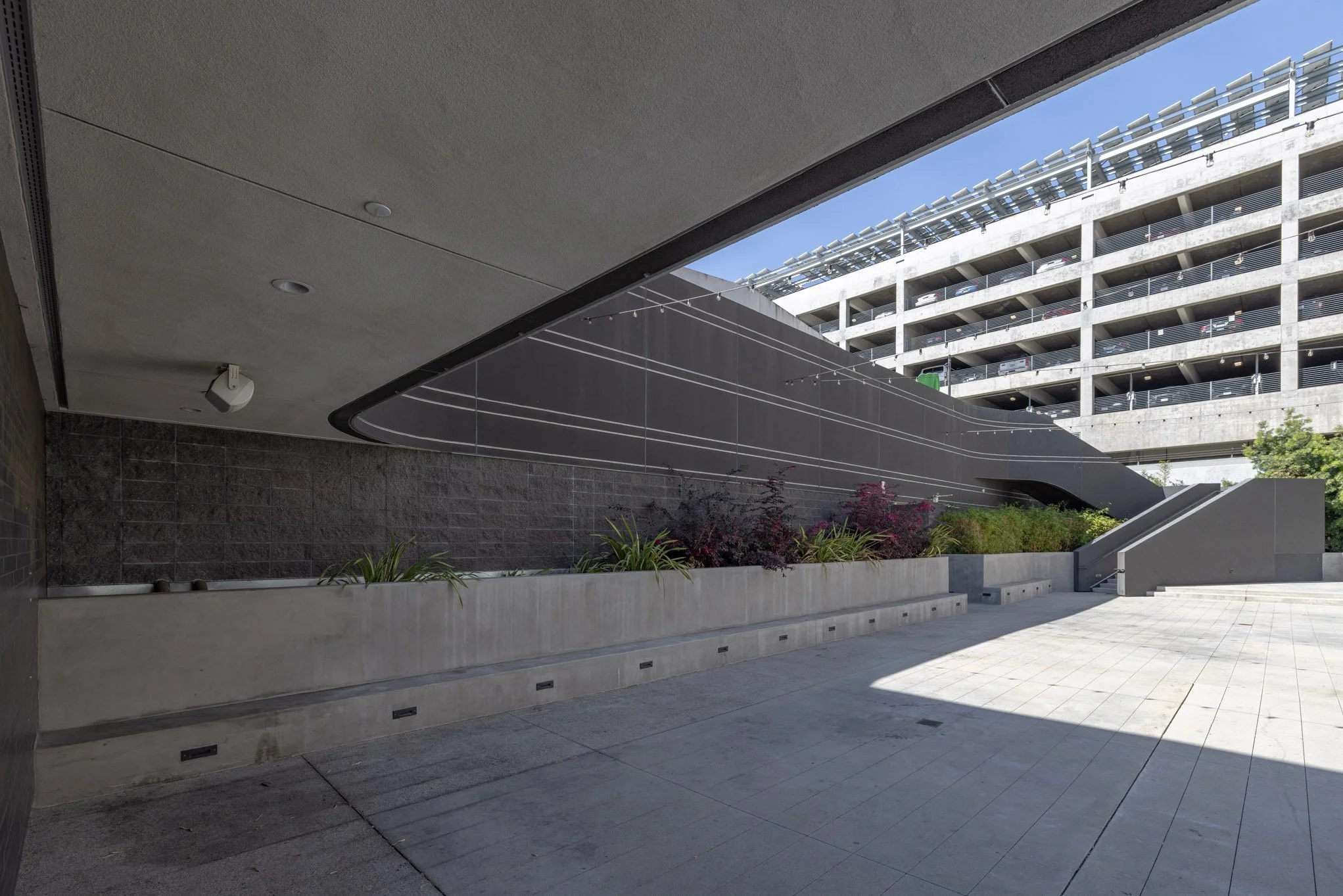 Urban outdoor area with a concrete staircase and parking structure in the background, green plants along the wall, and sunlight casting shadows on the tiled ground.