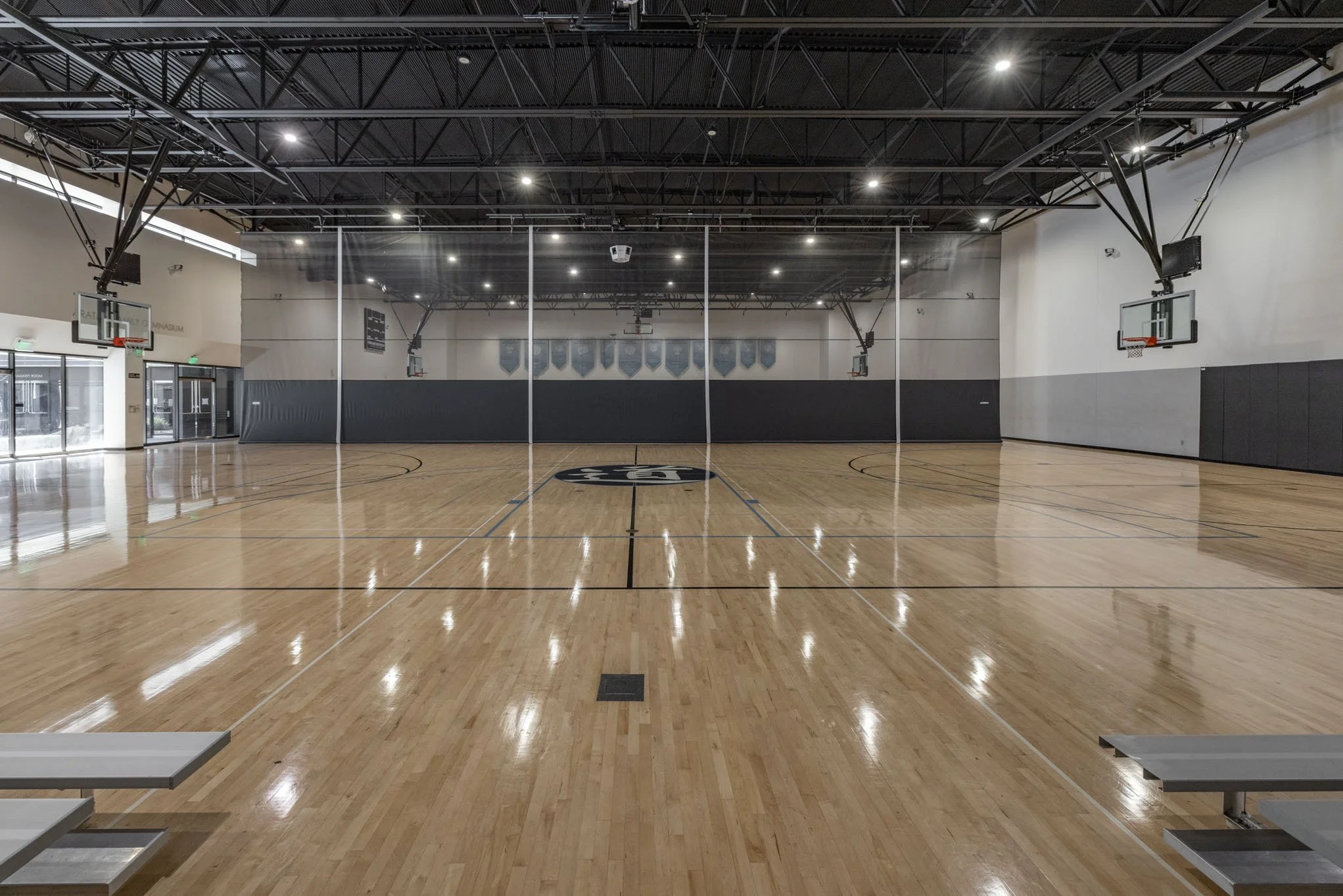 Empty indoor basketball court with polished wooden floor, basketball hoops, and a protective glass barrier in the center.