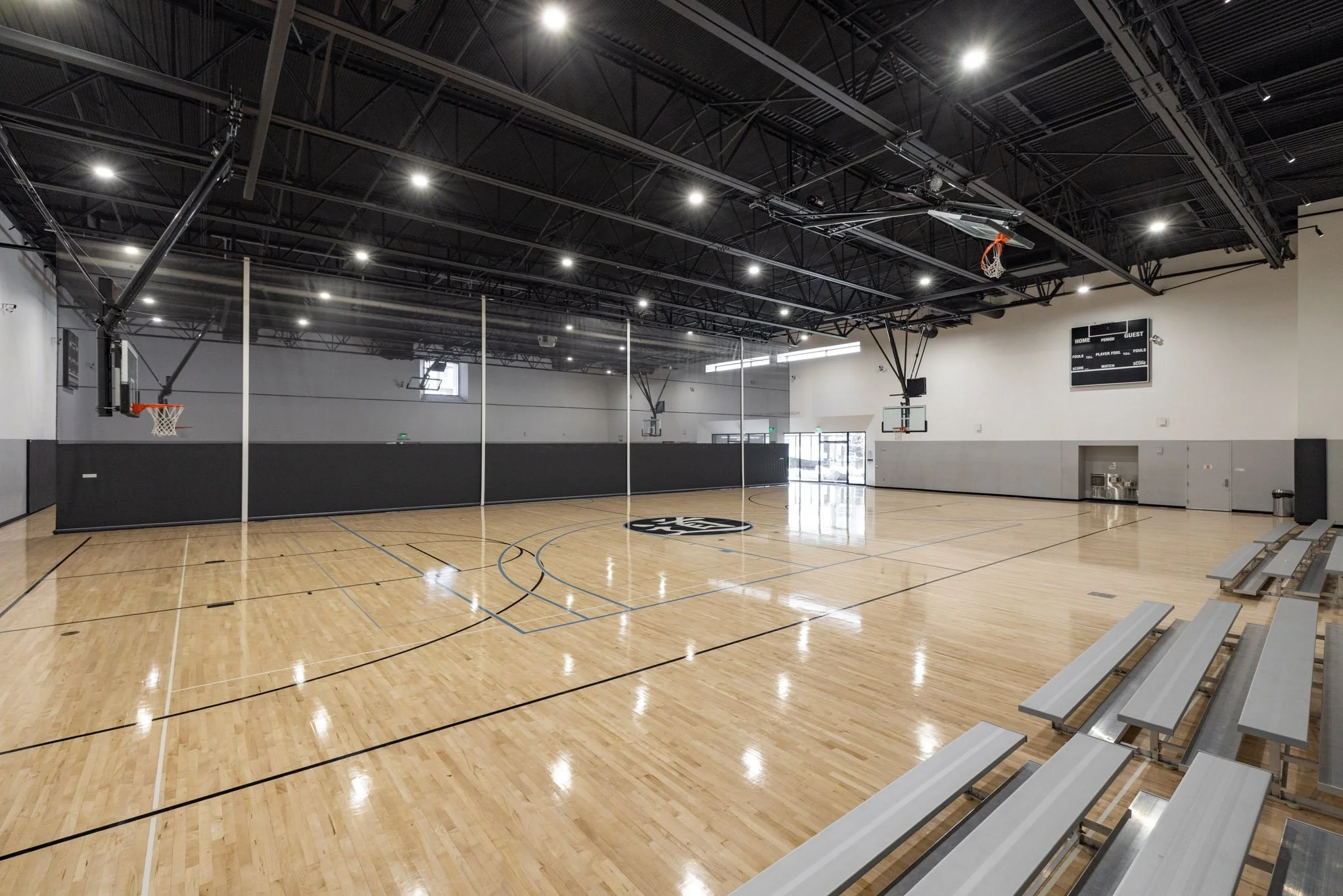 An empty indoor basketball court with a wooden floor, two basketball hoops, and a glass divider, inside a modern gymnasium with ceiling lights, bleachers on the side, and a digital scoreboard.