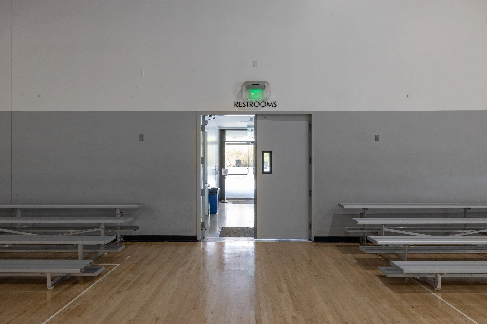 Empty gymnasium with metal benches on a wooden floor, view through open door to outside, sign for restrooms above door, exit sign illuminated above doorway.