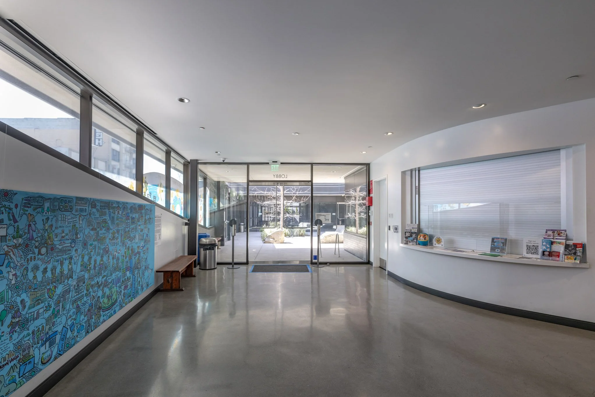 Interior of a modern building lobby with glass entrance doors, a curved white wall with a display kiosk, a bench, and large windows letting in natural light.
