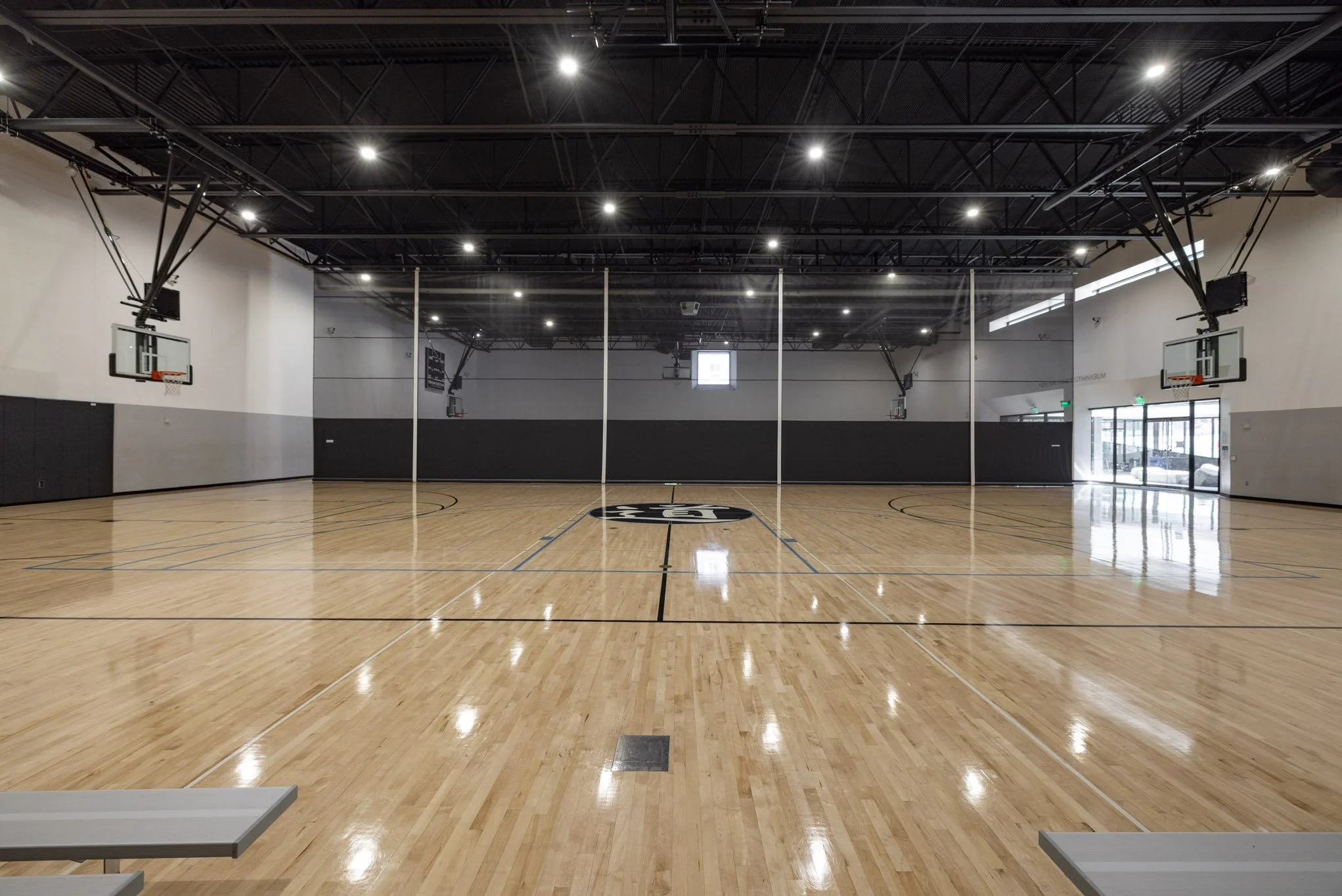 Empty indoor basketball court with polished wooden floor, black and white walls, multiple basketball hoops, and bright ceiling lights.