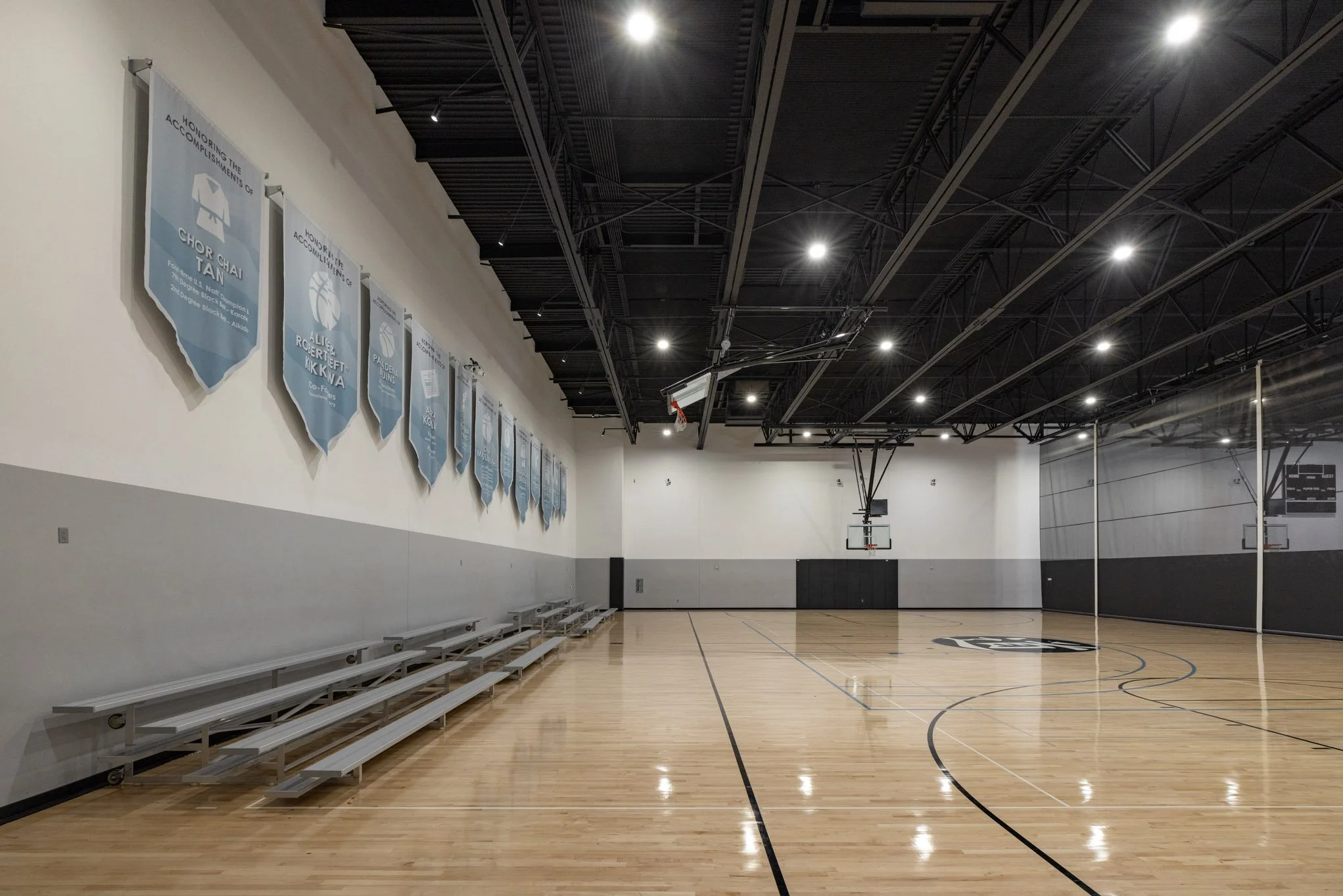 Empty indoor basketball court with polished wooden floor, black wall padding, basketball hoops, and blue banners hanging on the wall.
