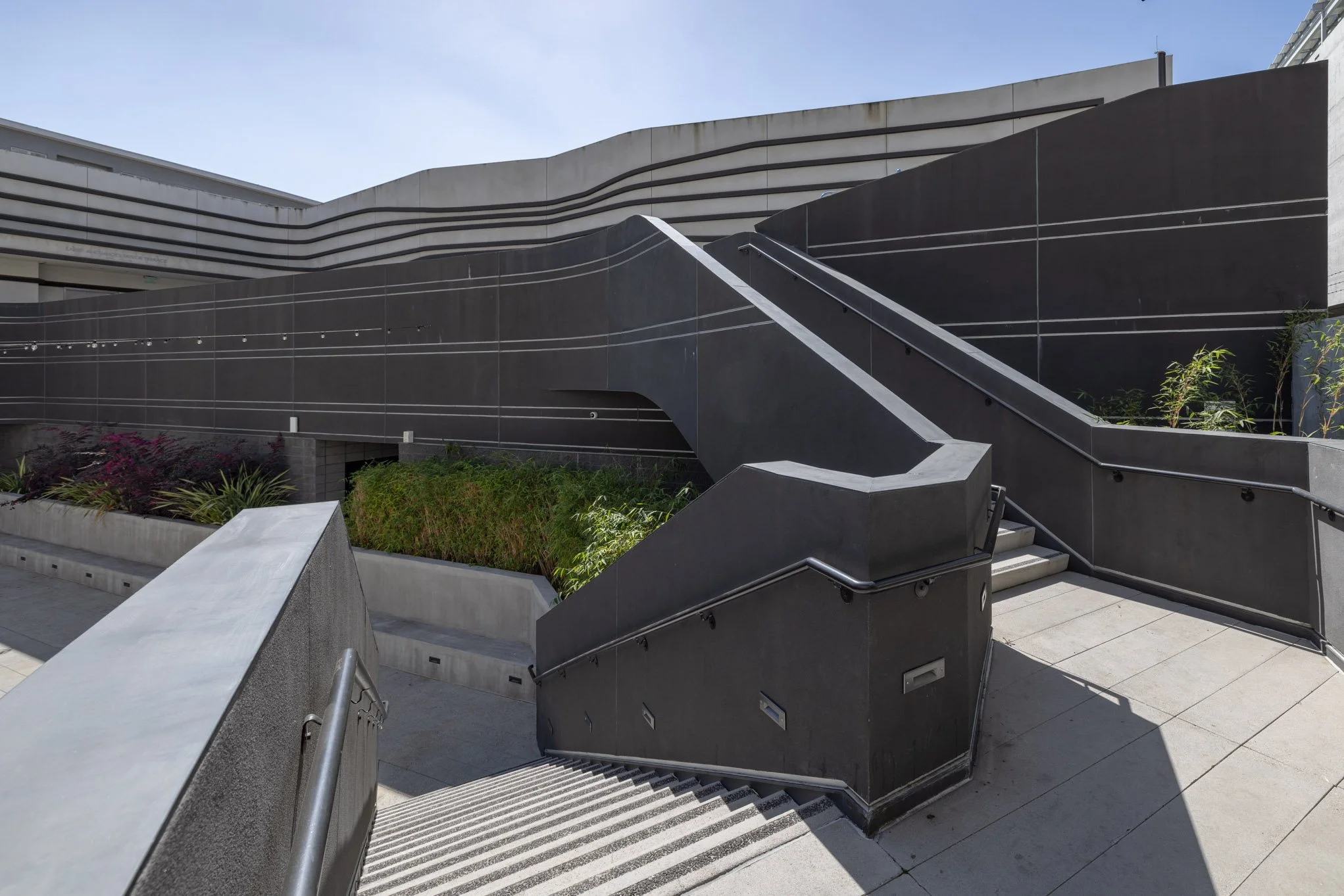 Modern urban outdoor staircase with black walls, metal handrails, and landscaped greenery underneath, part of a contemporary building exterior.