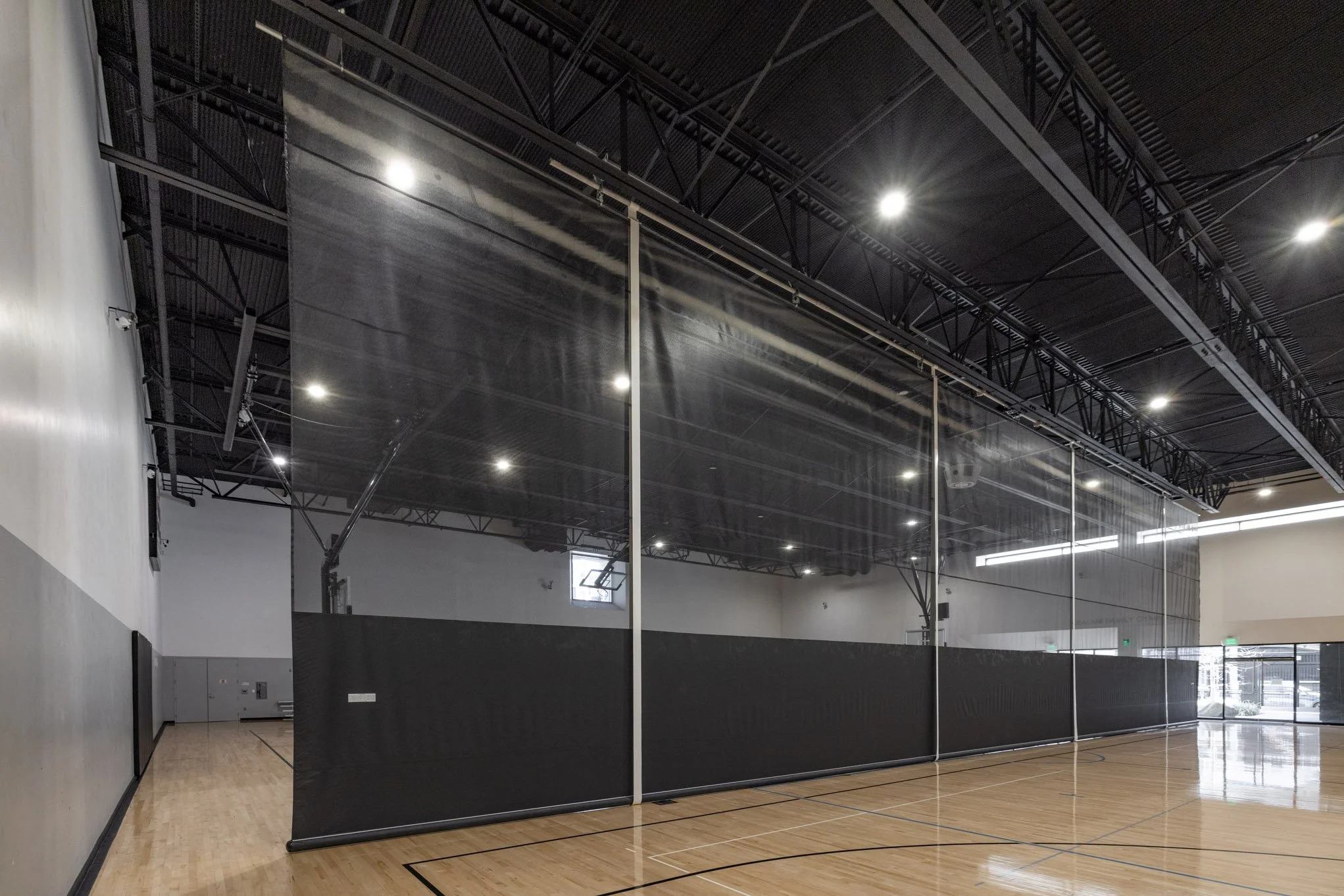 Indoor sports facility with black partition walls and high ceiling, illuminated by ceiling lights, and a polished wooden floor.