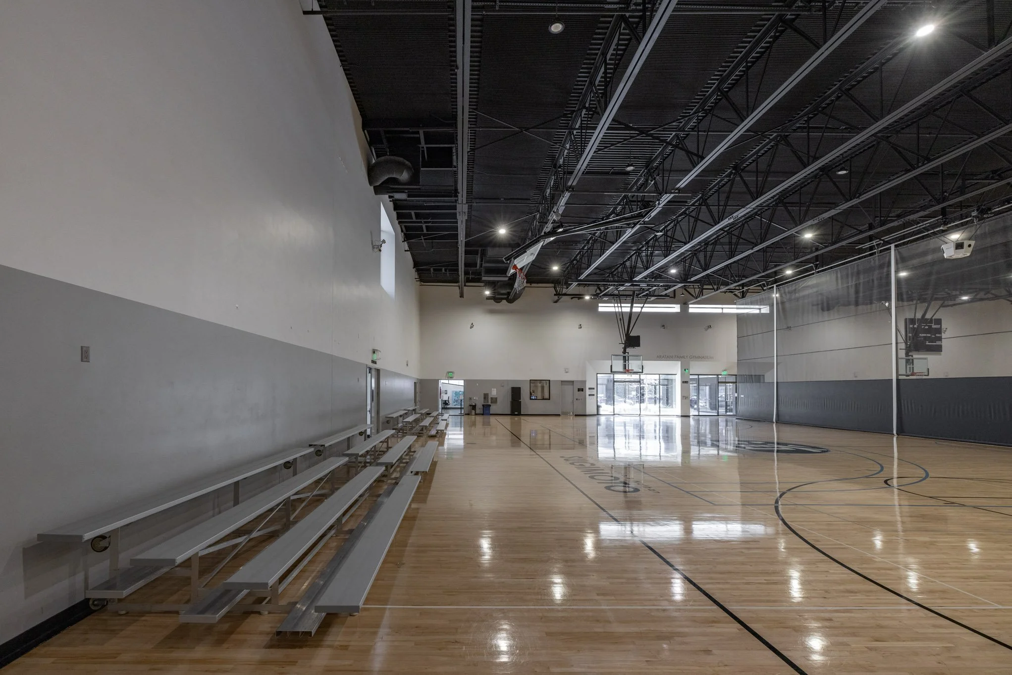 Empty indoor basketball gymnasium with polished wooden floor, high ceiling with black beams, and basketball hoops at each end. Bleachers are pushed against the wall on the left side, and glass doors leading outside are at the far end.