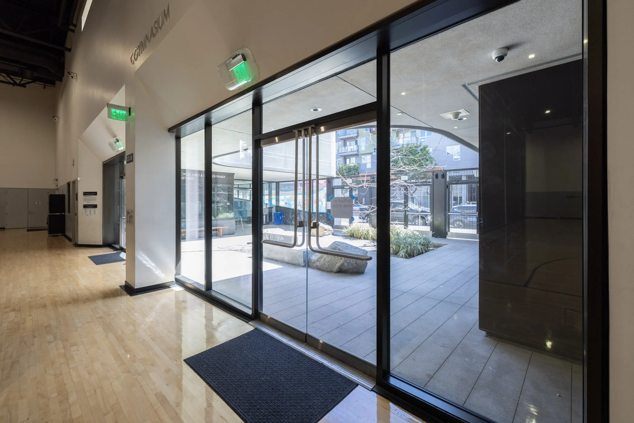 Glass entrance door with an outdoor courtyard visible through the glass, inside has wooden flooring and elevator to the left.