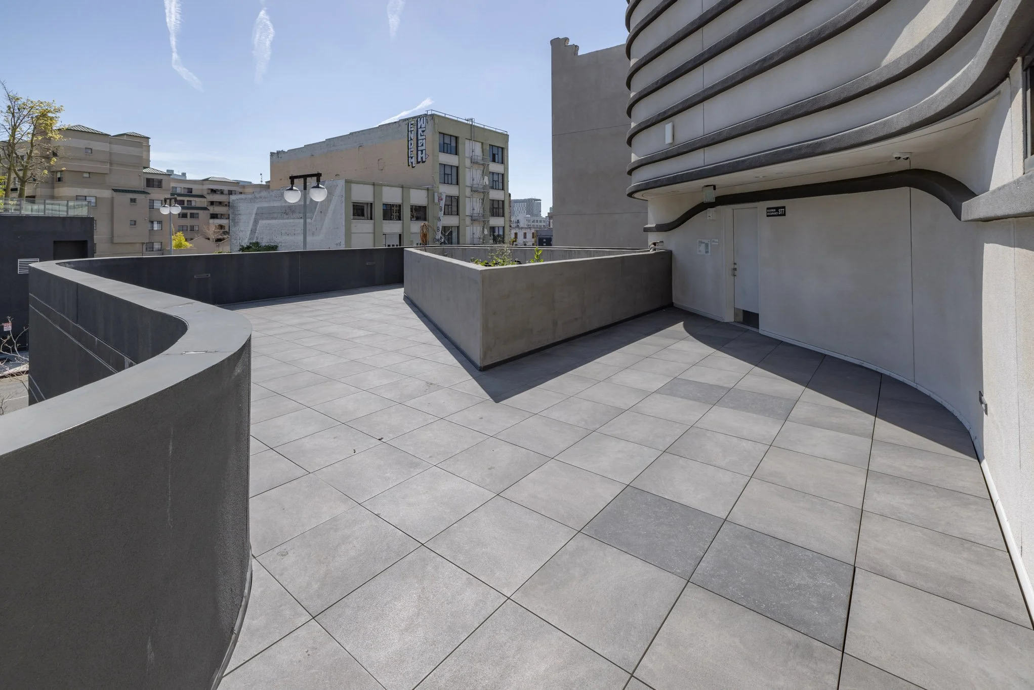 Empty rooftop terrace with gray tiled floor, curved gray concrete walls, and a modern building exterior with neighboring buildings visible in the background under a clear blue sky.