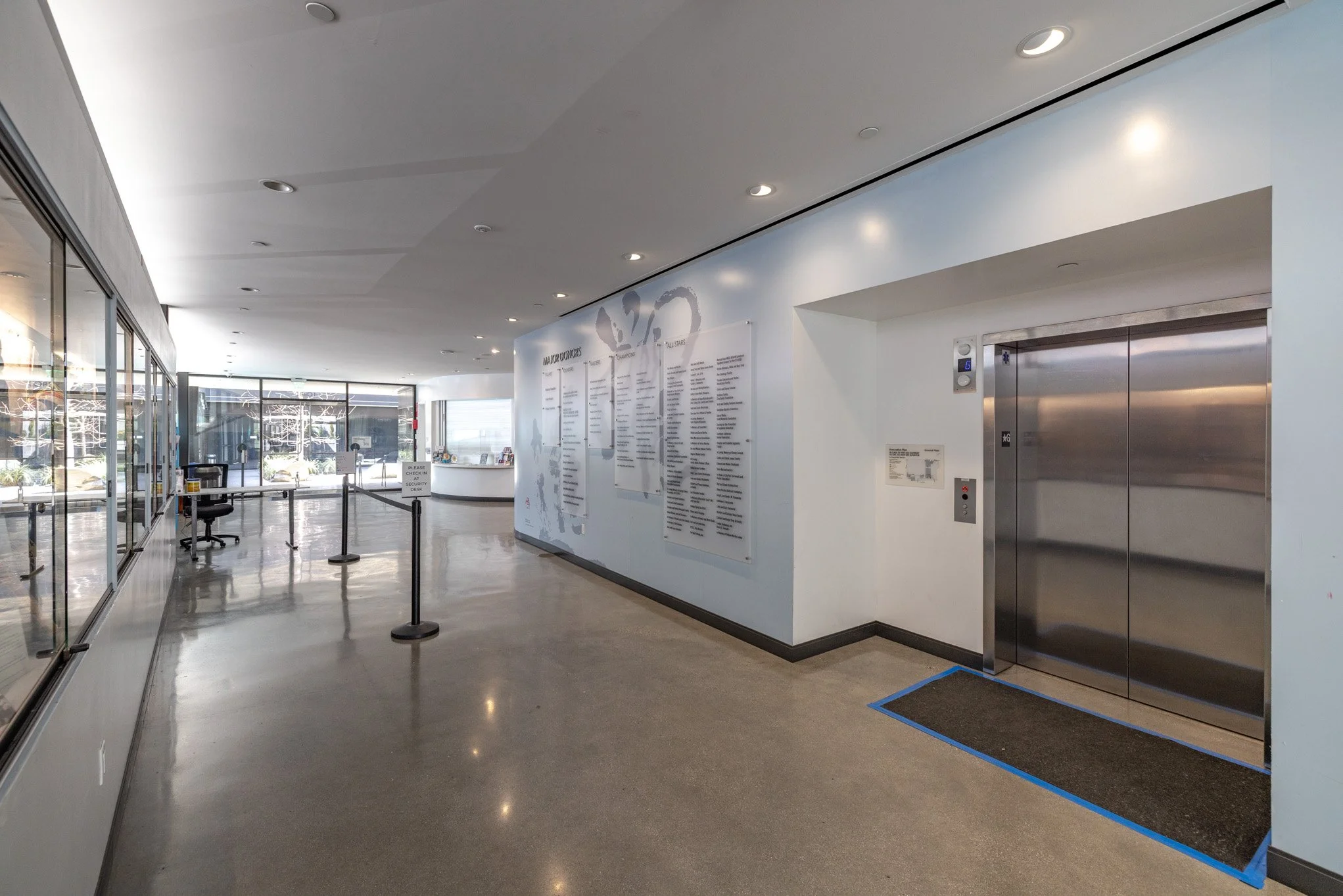 Lobby area with elevator, reception desk, informational boards on the wall, and glass walls leading outside.