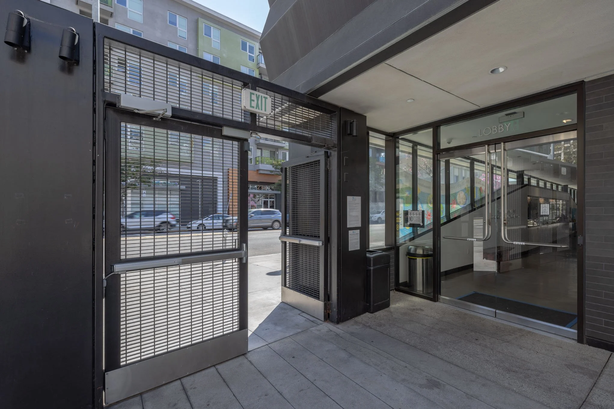 Entrance to a building with a glass door marked 'Lobby' and a metal gate with an exit sign above, located on a city street.