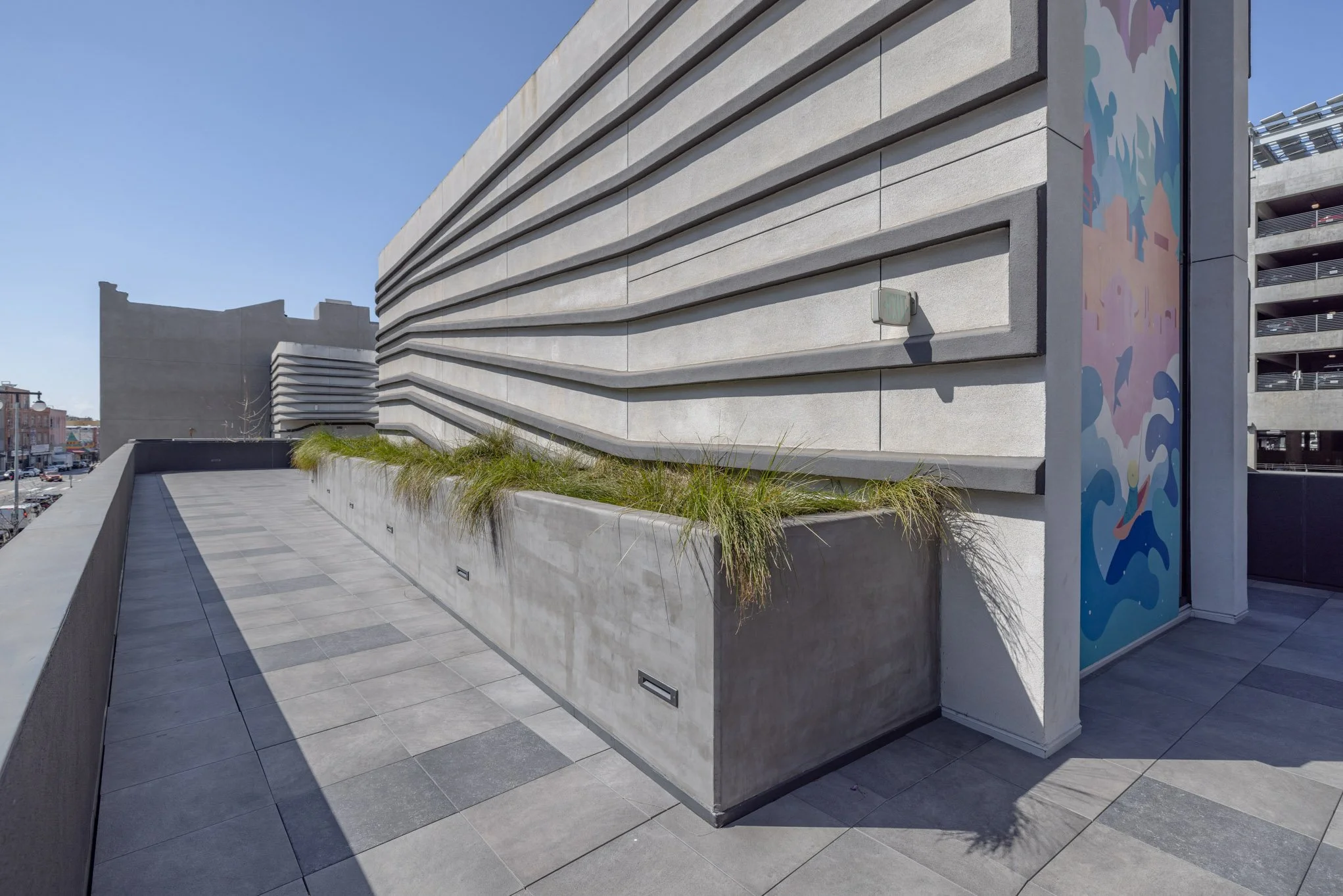 Modern rooftop terrace with concrete planter filled with grass, adjacent to a contemporary building with textured horizontal concrete panels and a colorful mural.